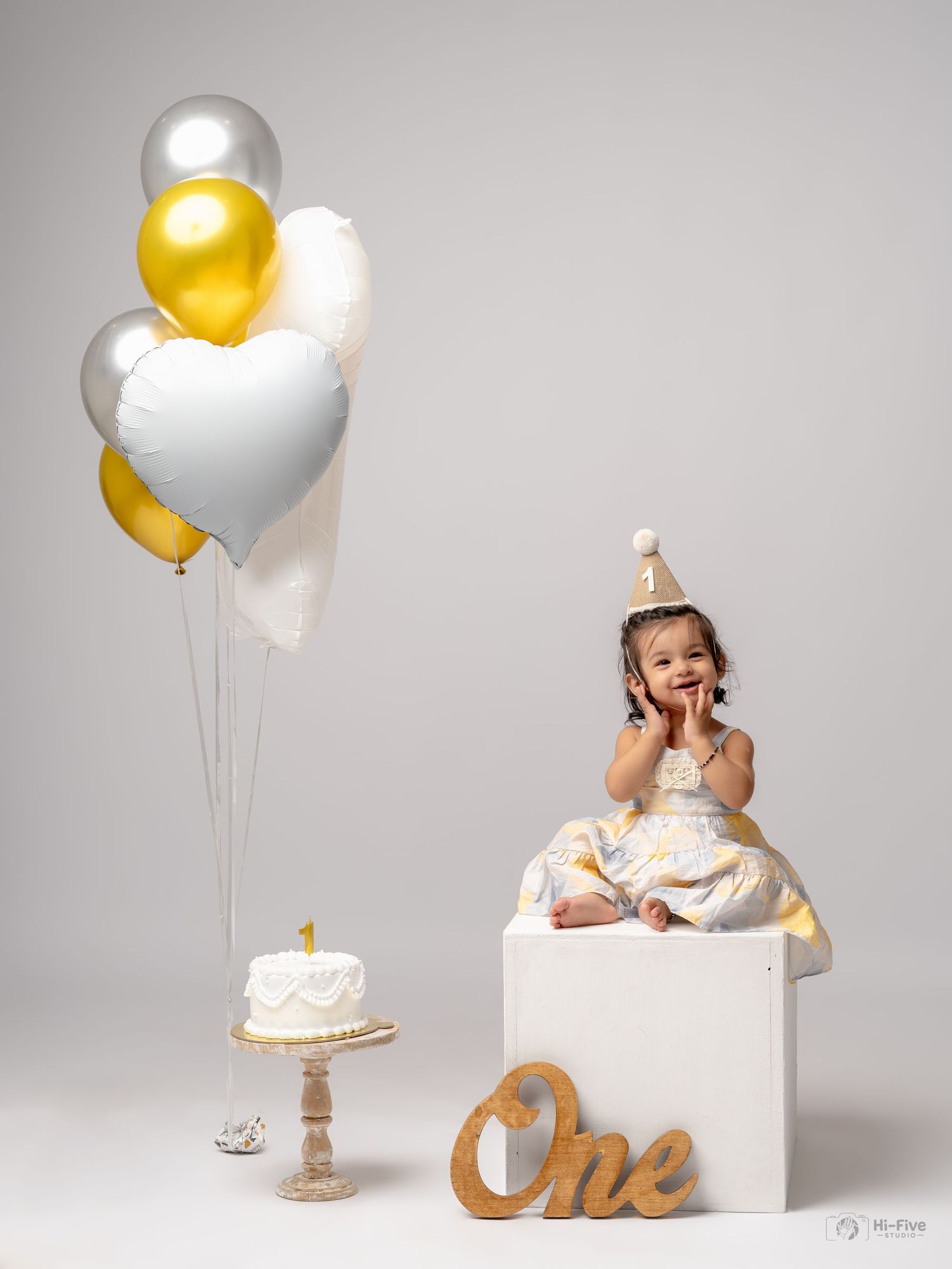 Baby girl in white gold dress celebrates first birthday with balloons and smash cake in studio photo.