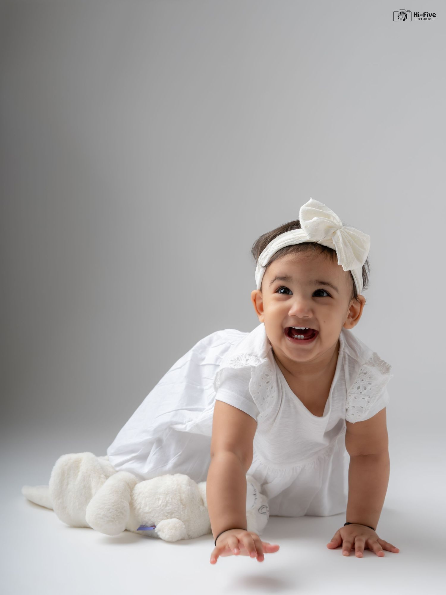 Smiling baby girl in white dress and bow headband crawling on floor with stuffed bunny toy.