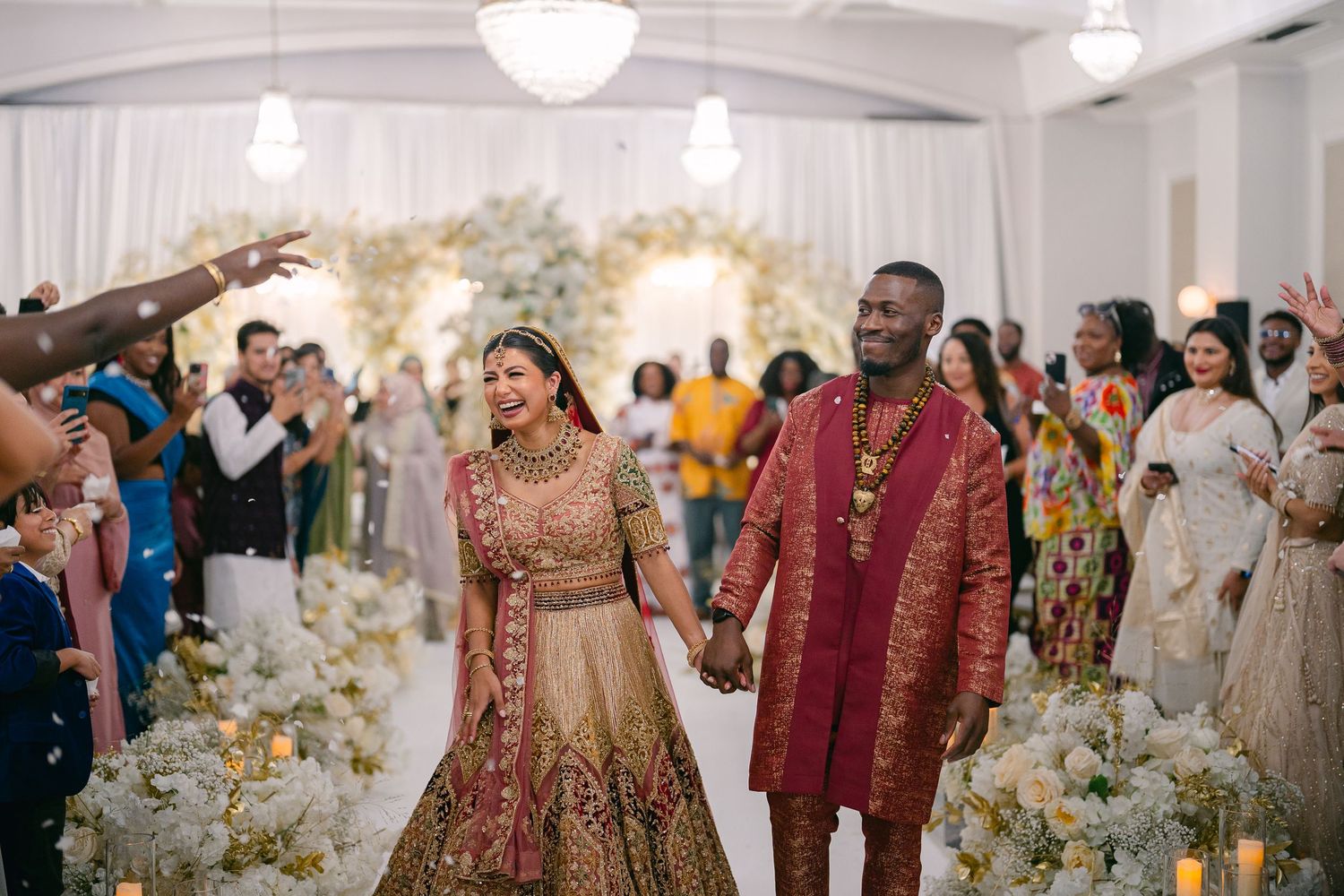 Guests in traditional Indian wedding attire dance and celebrate at an elegant reception decorated with white floral arrangements.