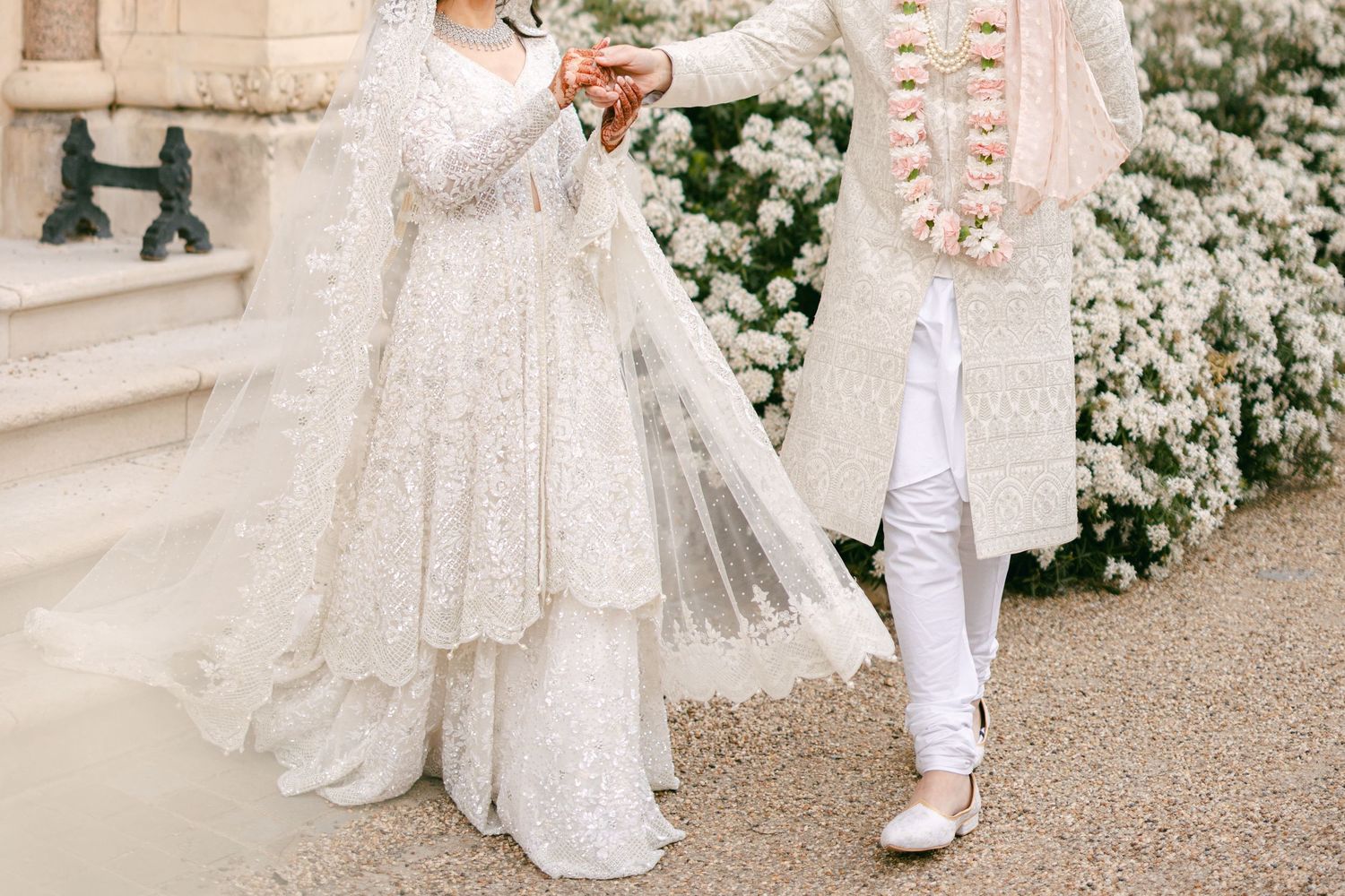 South Asian couple in white wedding attire holding hands, bride in embroidered lehenga with veil, groom in sherwani with floral garland.