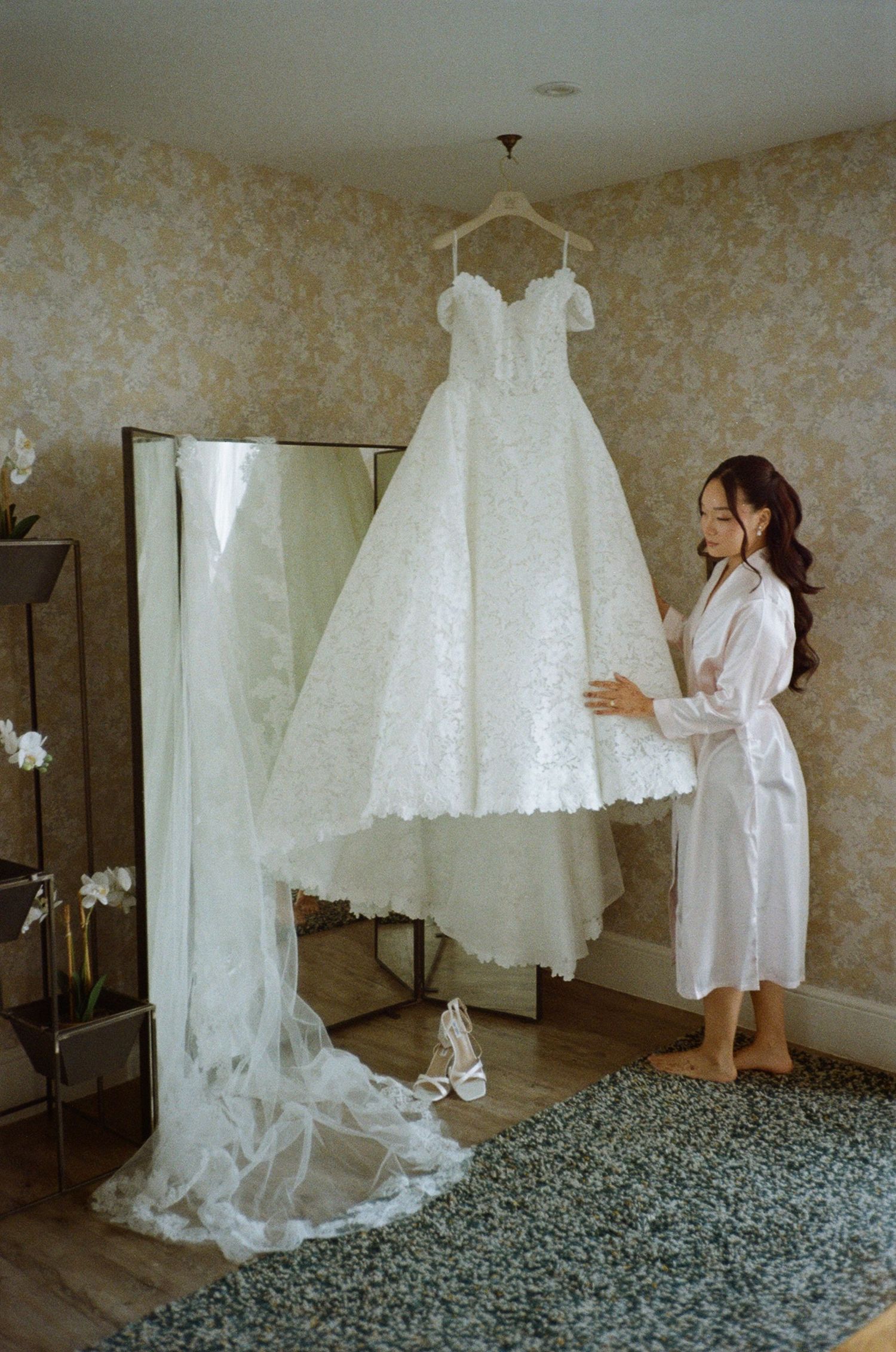 Bride in white robe admiring her lace wedding gown hanging in a floral wallpapered bridal suite.