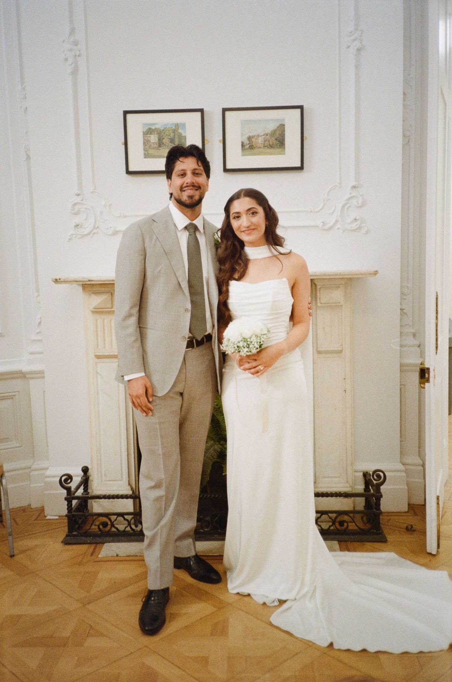 Bride and groom posing together in elegant white room, she in white gown with bouquet, he in tan suit with green tie.
