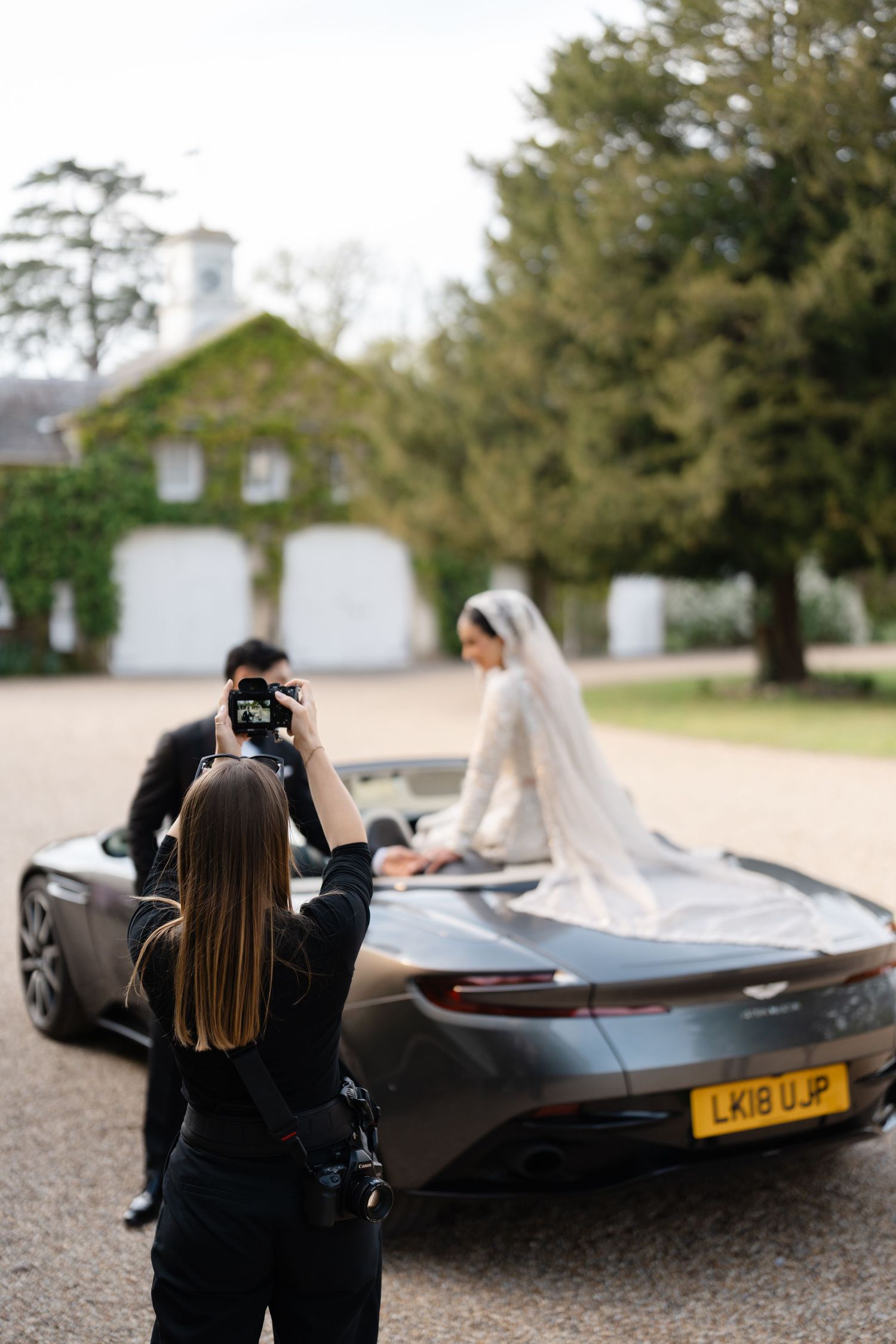 Photographer capturing bride in wedding dress posing on Aston Martin DB11 at elegant UK countryside venue.