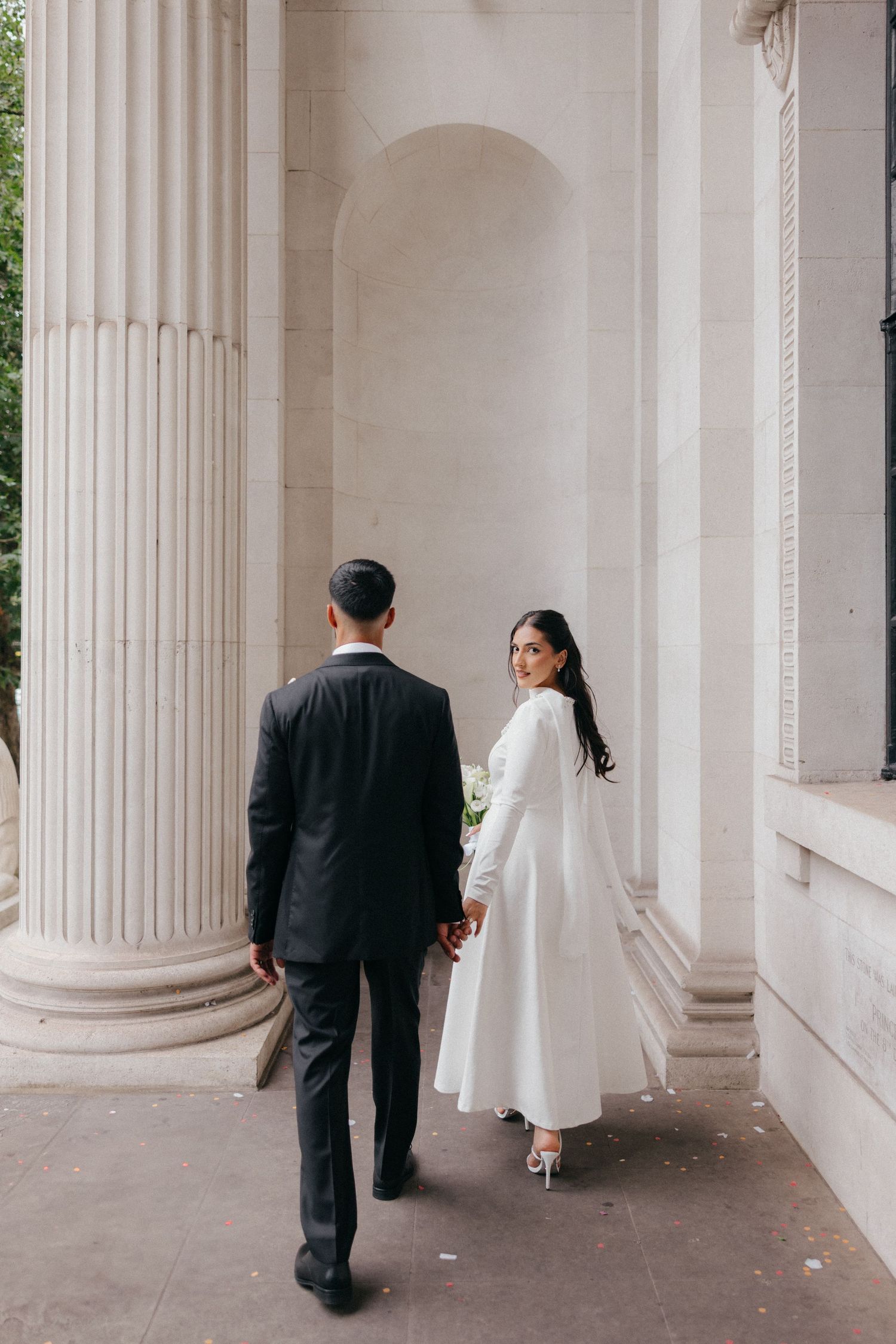 A wedding couple walks through an elegant colonnade with white pillars.