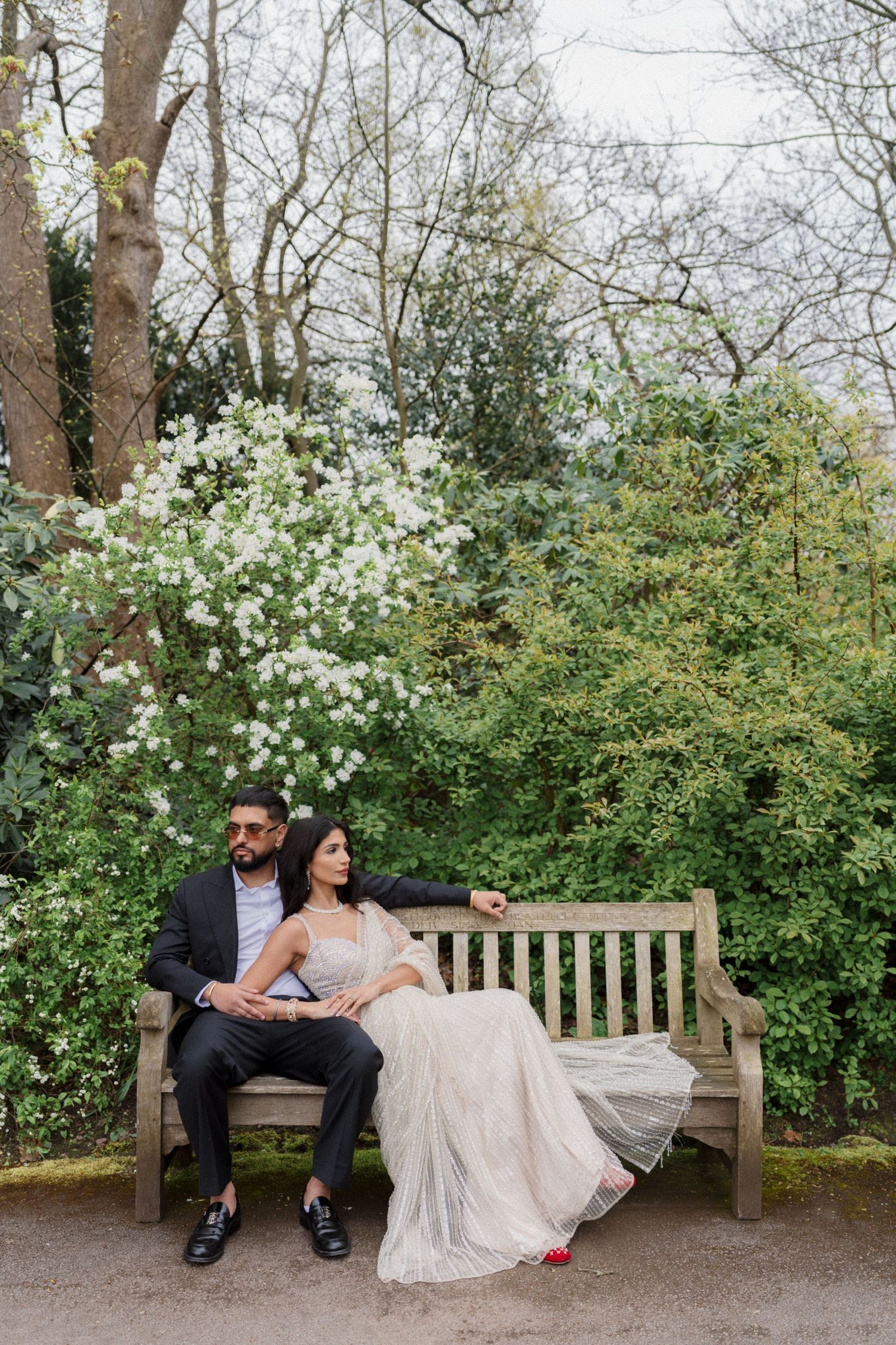 Couple in wedding attire sitting on a wooden bench surrounded by lush greenery and white blossoms in a garden.
