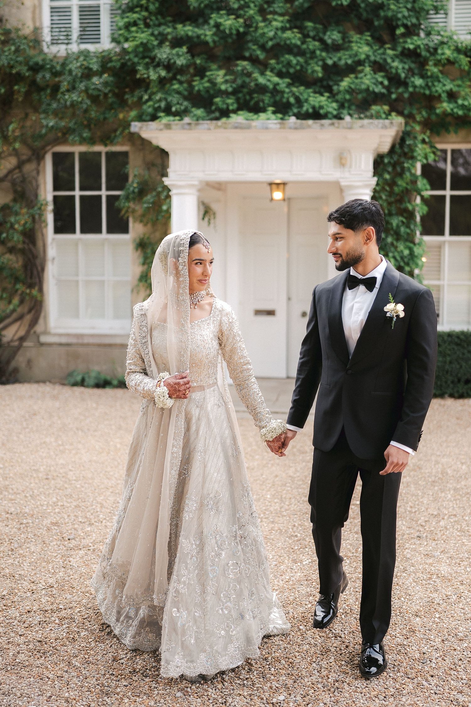 Bride in embellished white lehenga with veil holds hands with groom in black tuxedo outside elegant manor.