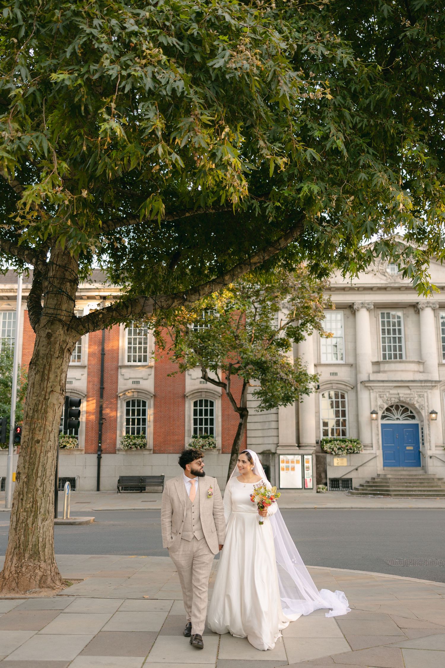 A wedding couple poses under trees in front of a historic building on their special day.