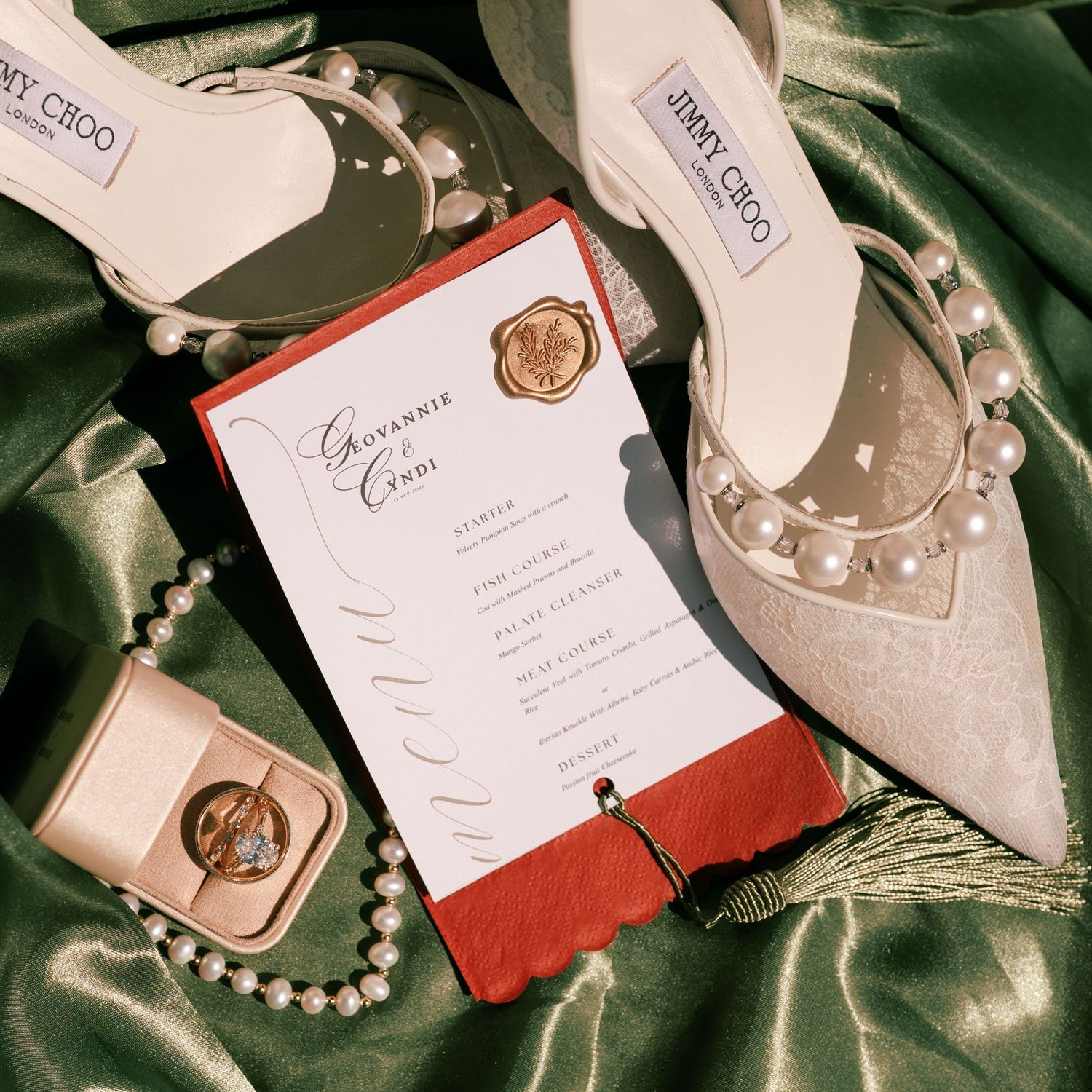 Elegant wedding flat lay with Jimmy Choo heels, pearl necklace, wax seal menu card, and gold ring box on green silk.
