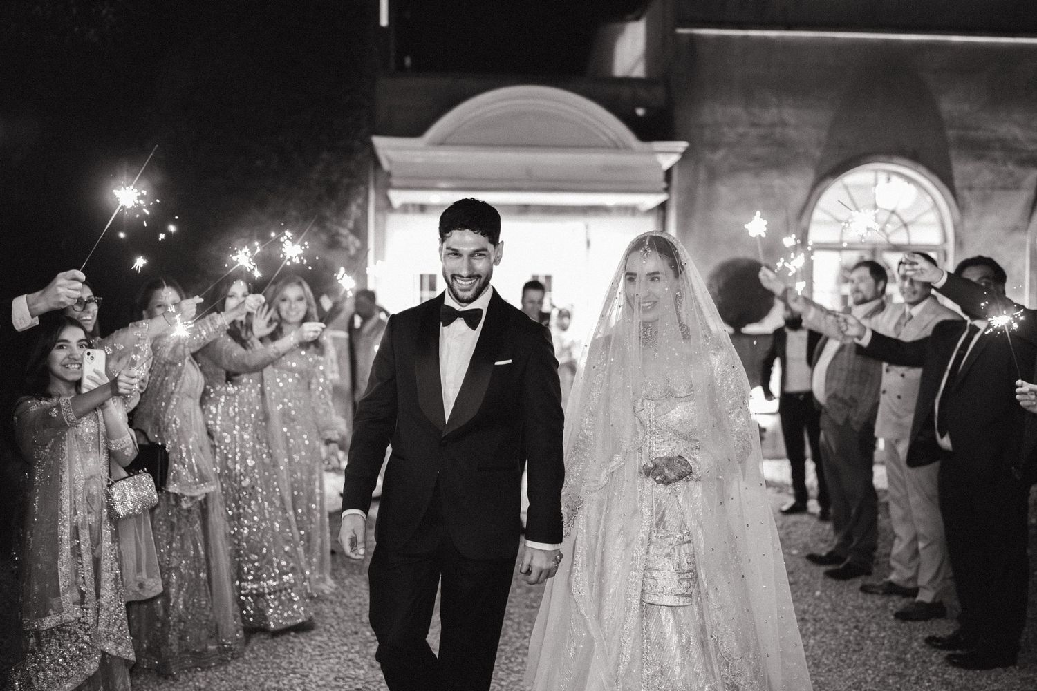 Black and white wedding photo of couple walking through sparkler send-off, bride in lace gown, groom in tuxedo.