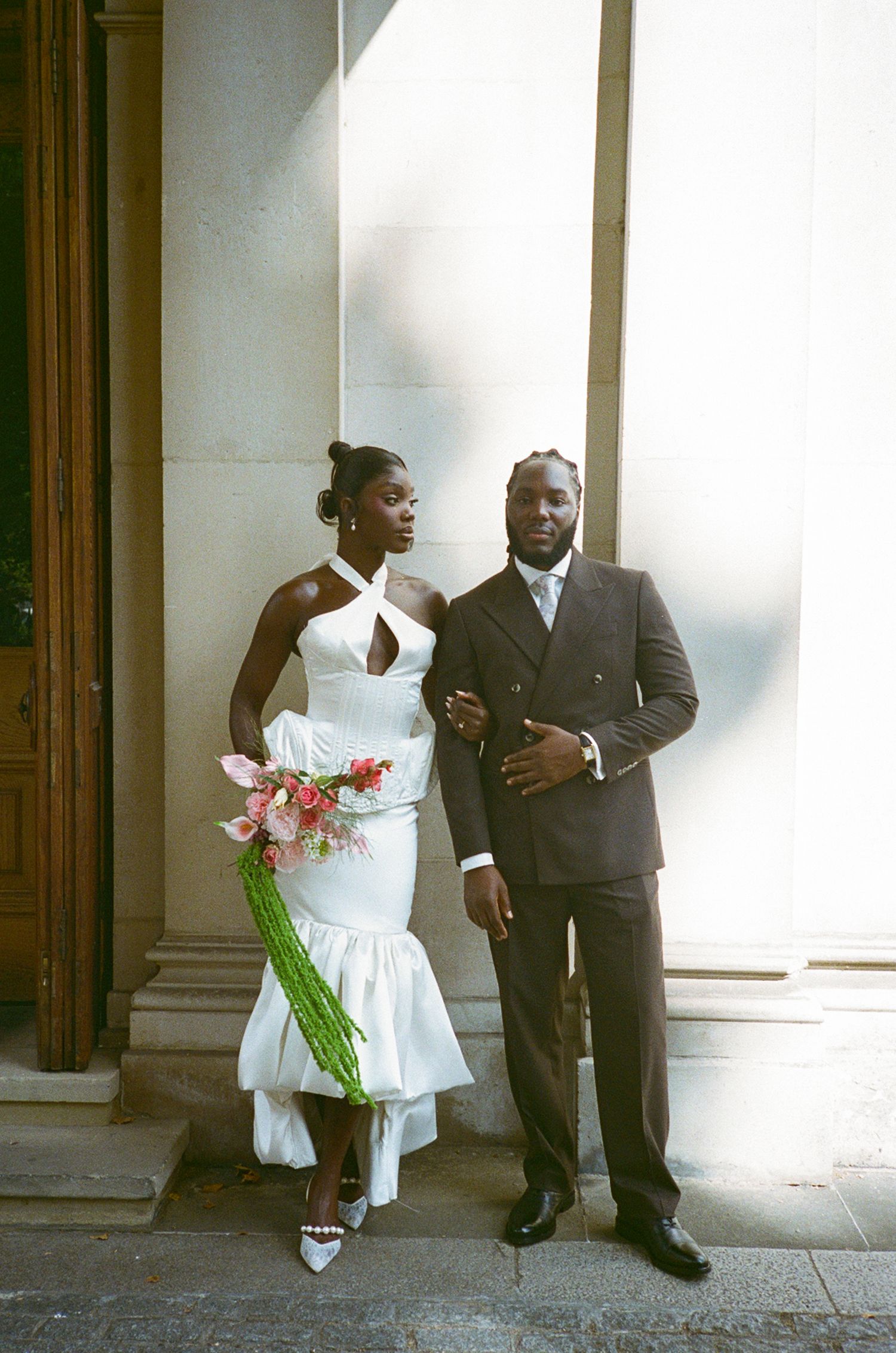 A bride in a white halter mermaid gown holds a pink bouquet beside a groom in a dark suit outside a building.