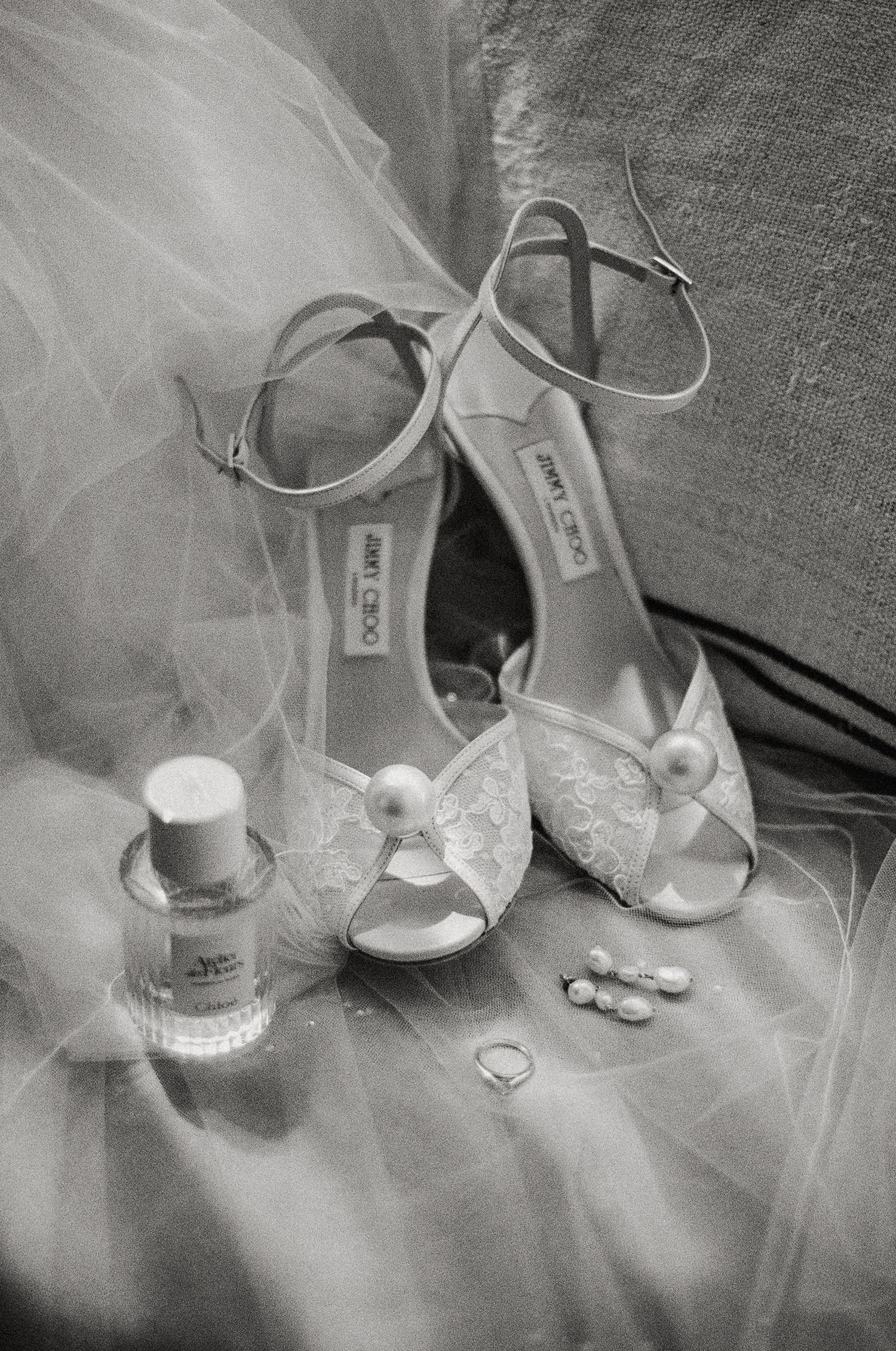 Black and white photo of Jimmy Choo bridal heels with pearl accents, perfume bottle, rings, and earrings on a wedding veil.