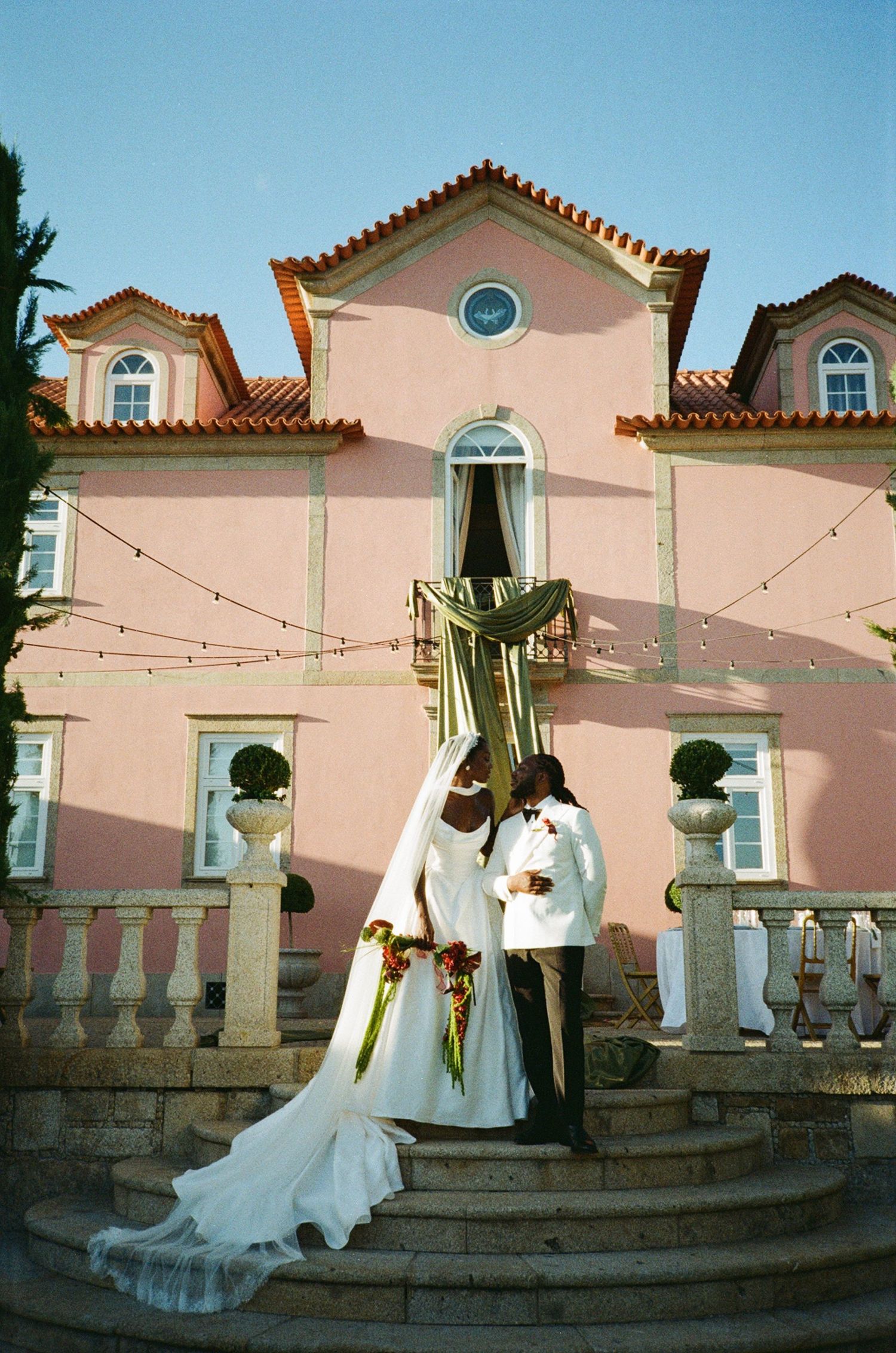 Bride and groom in elegant white attire pose on stone steps before a romantic pink villa with string lights.