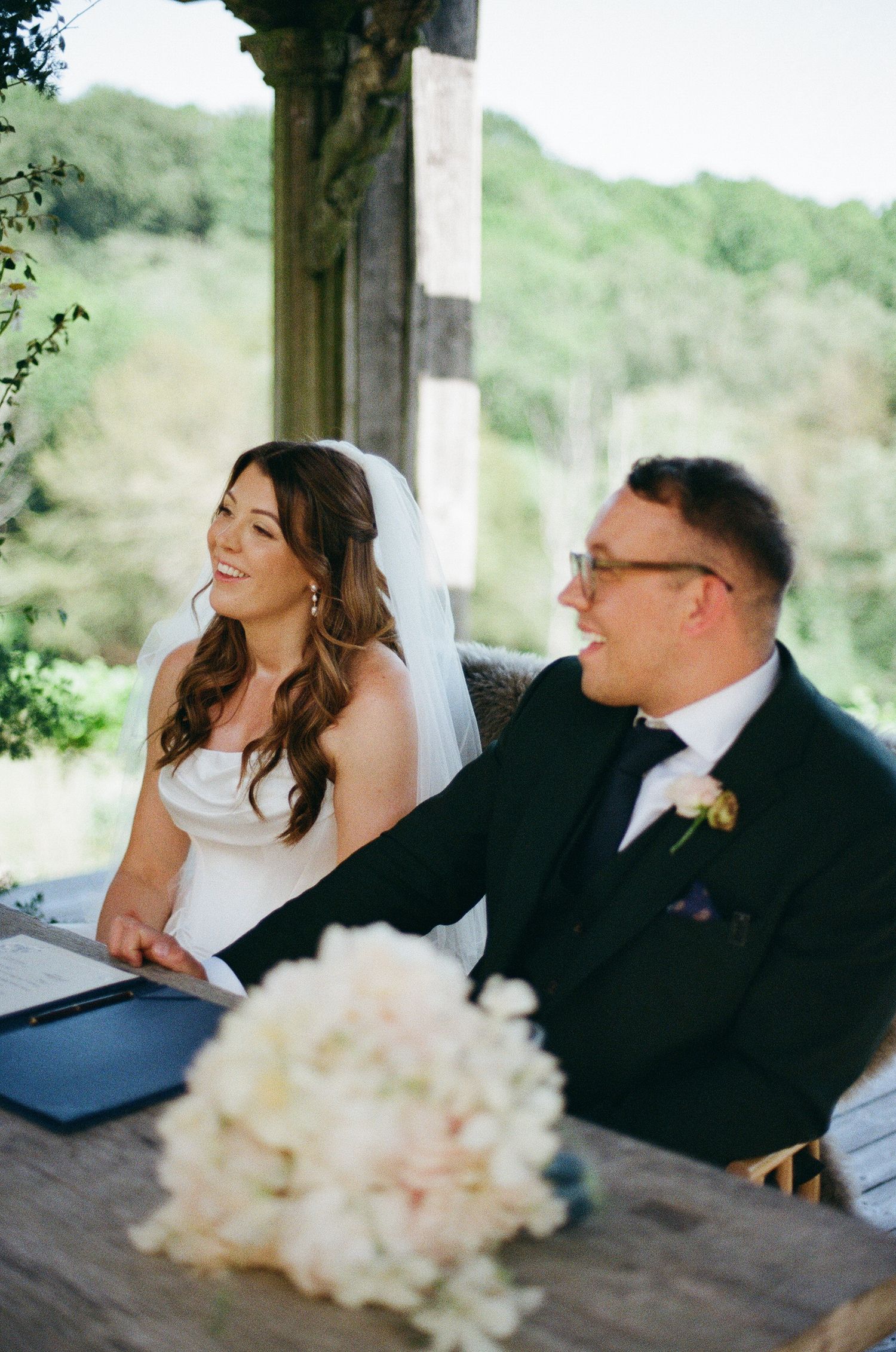 Bride and groom smiling at wedding table with white floral bouquet, lush green trees in background.