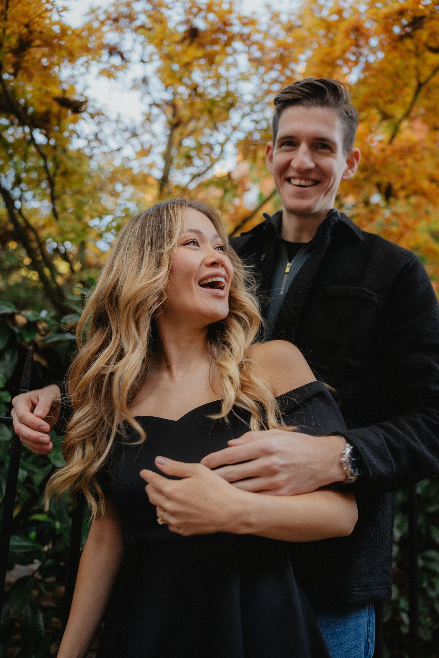 A couple laughs together against a backdrop of vibrant fall foliage.