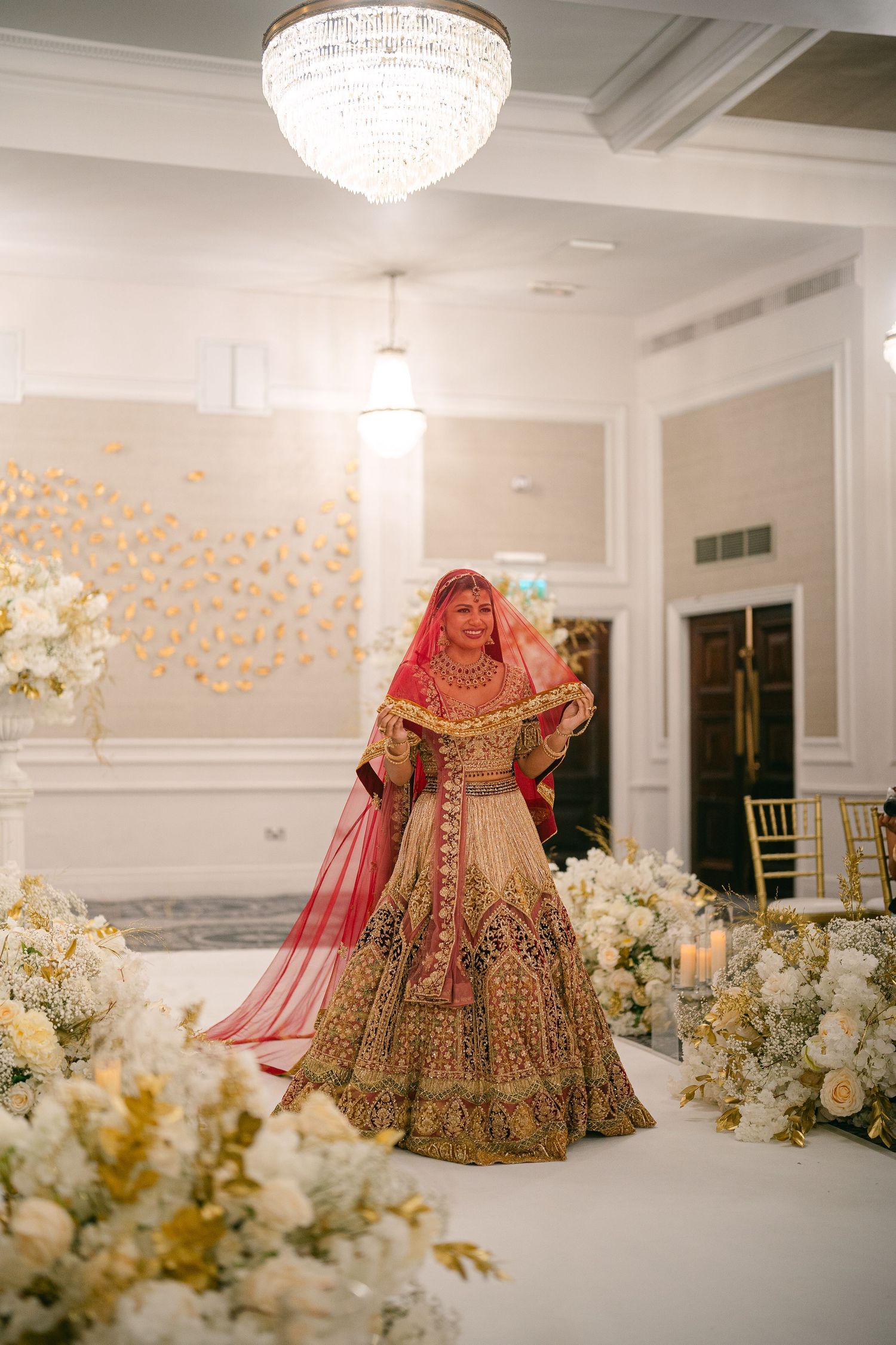 A bride in an ornate gold and red lehenga poses elegantly among white floral decorations under a crystal chandelier.