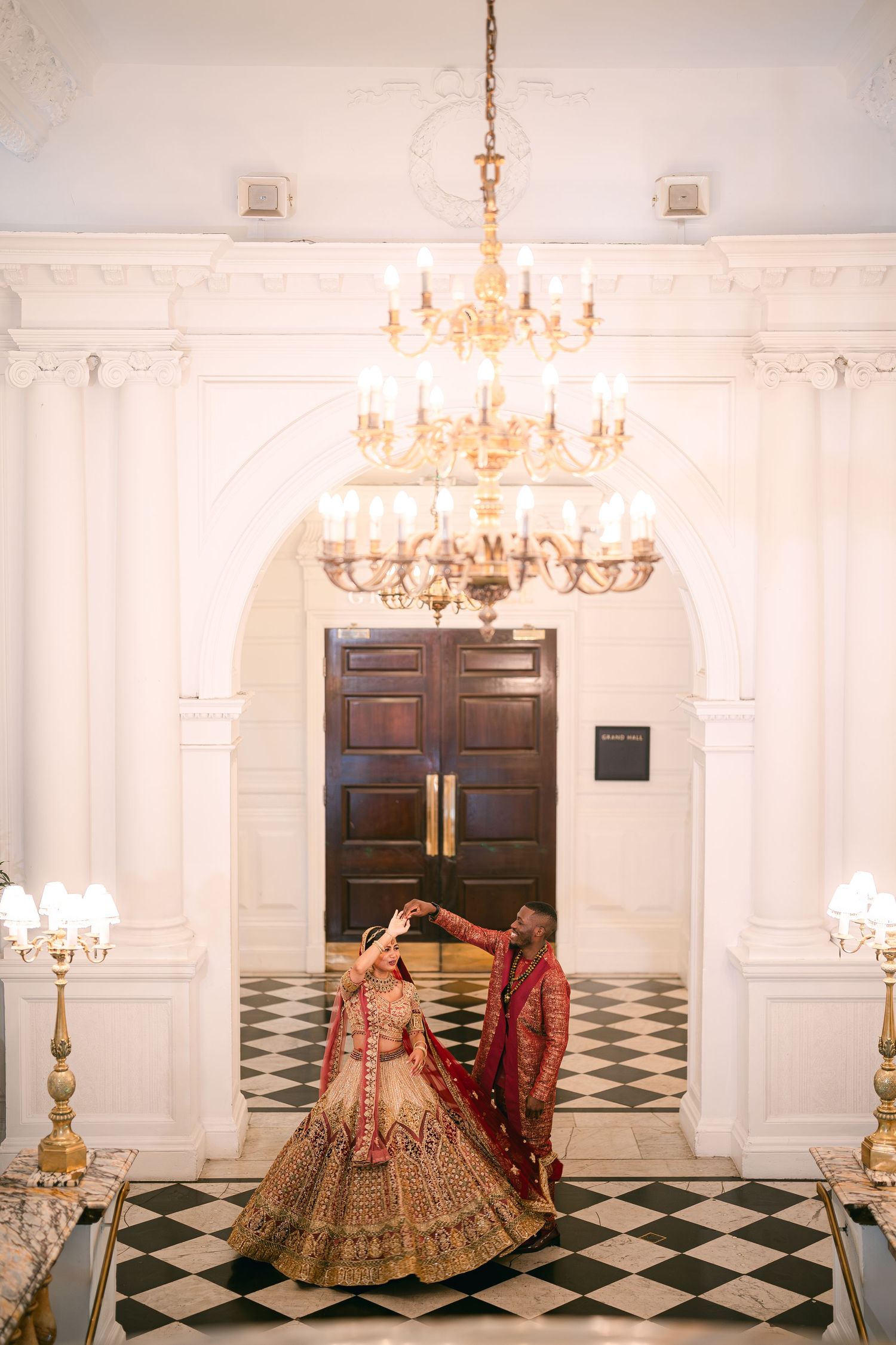 Elegant white venue with checkerboard floor and crystal chandeliers featuring traditional Indian bridal attire.