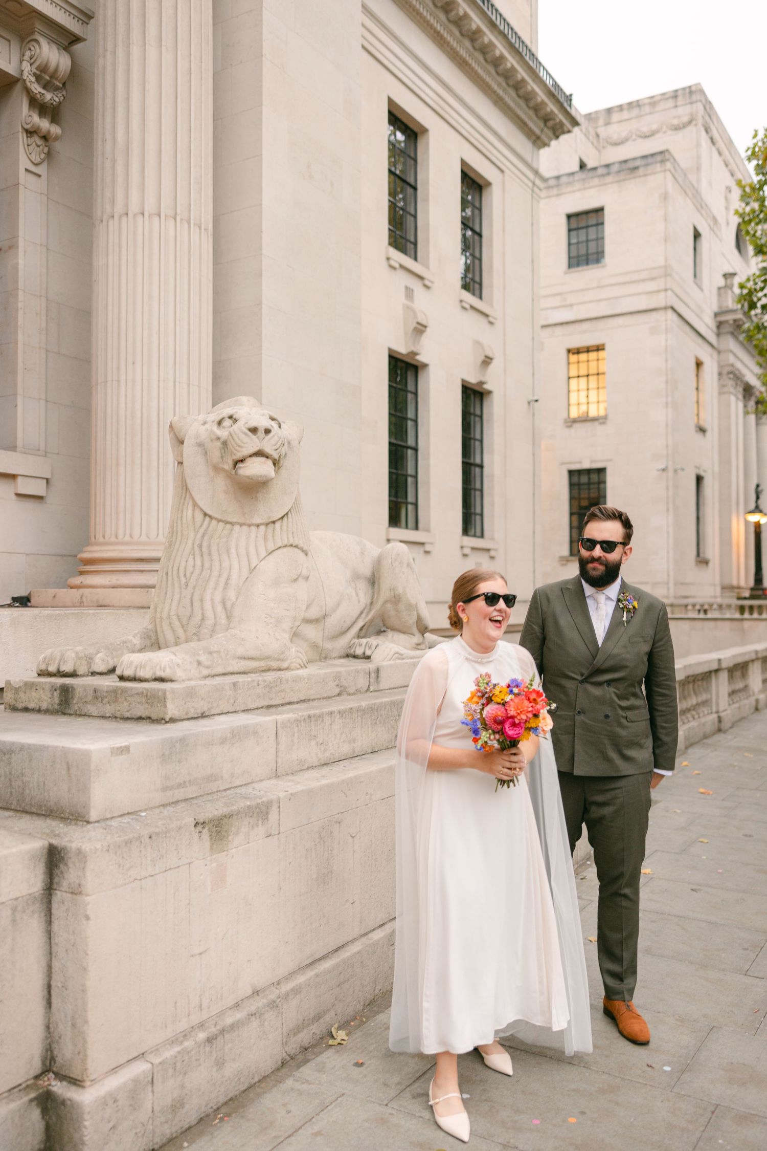 A couple poses with bright bouquet next to stone lion statue at historic building.