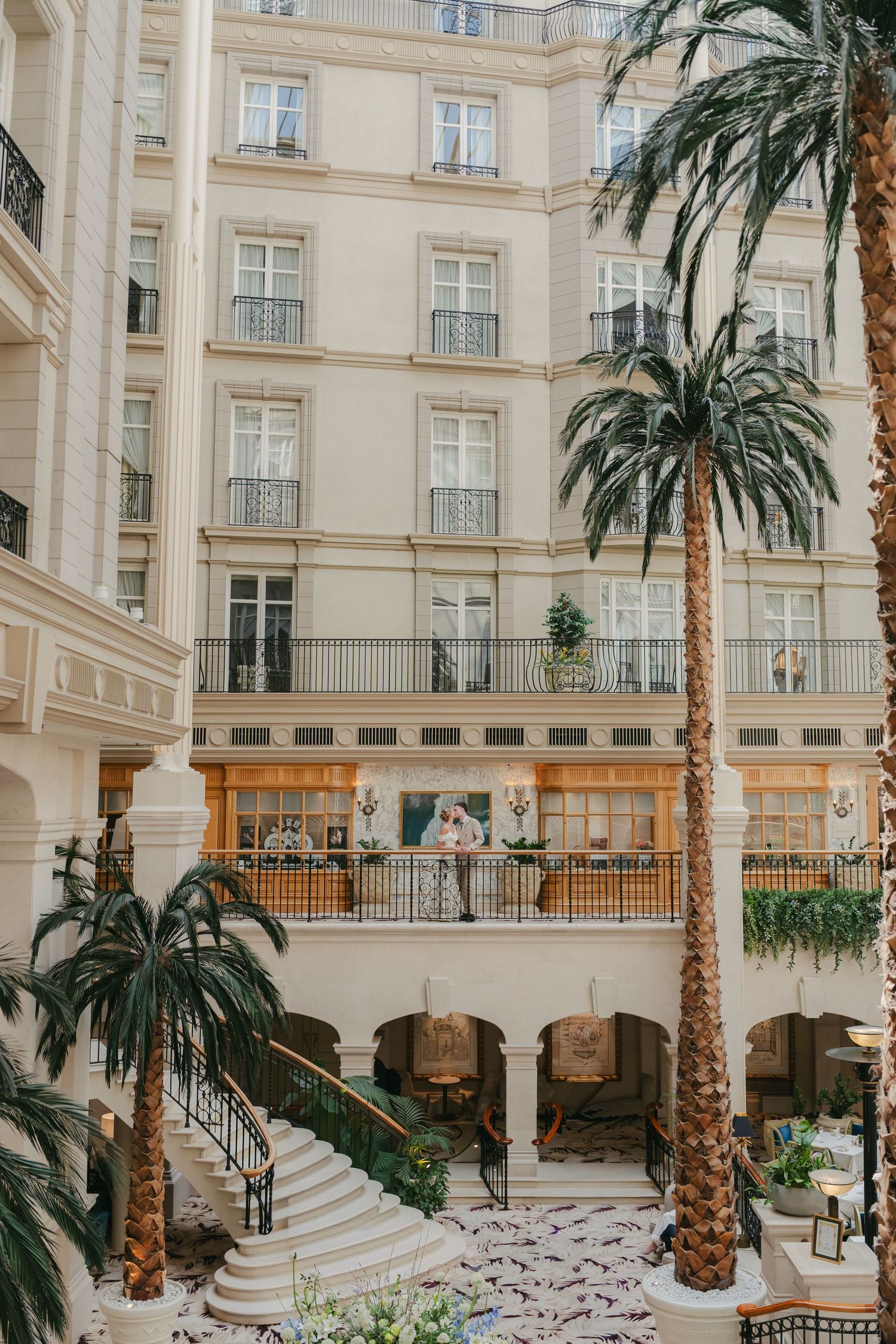 Elegant hotel atrium with tall palm trees, grand staircase, and Haussmann-style facade featuring ornate balconies.