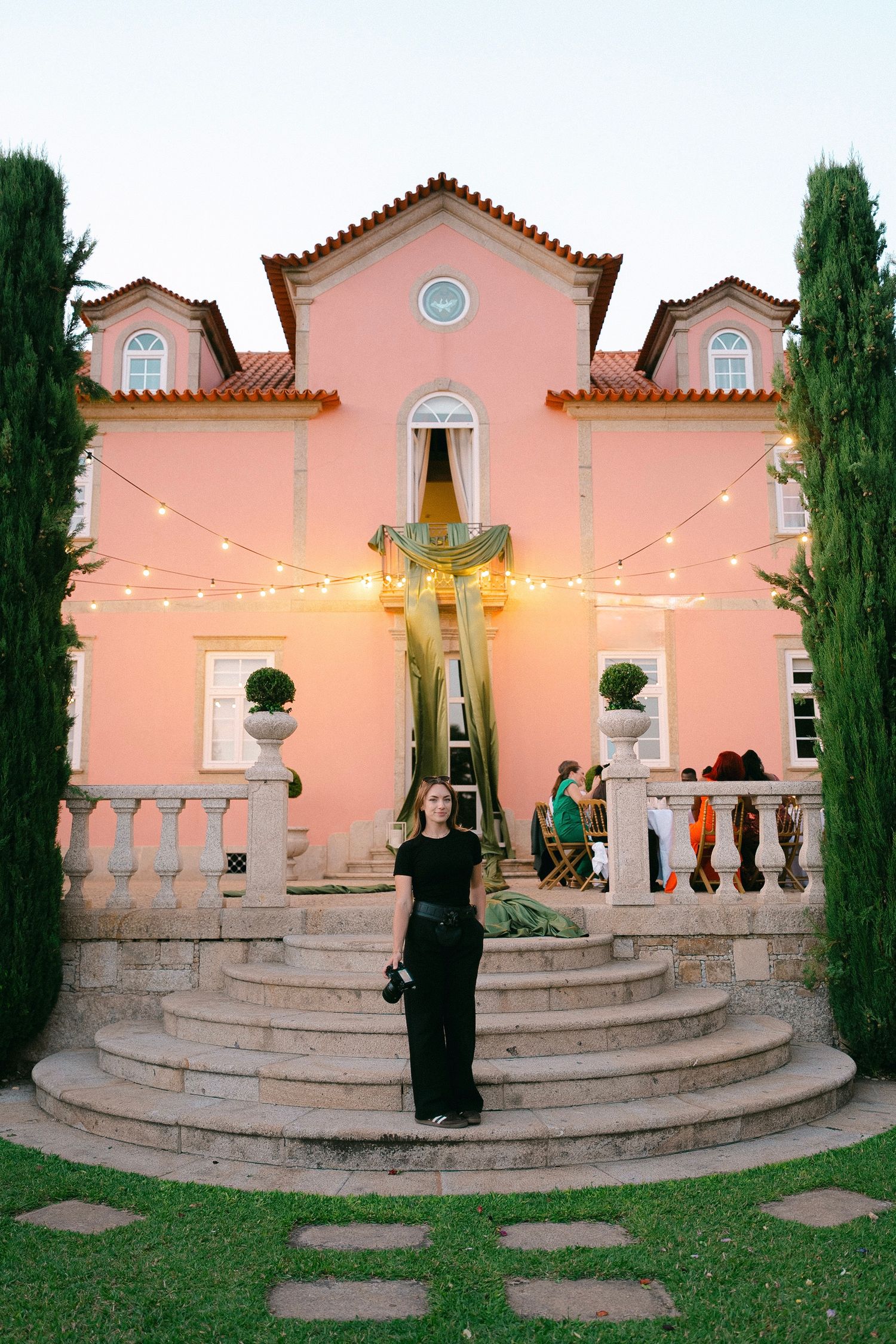 Woman in black outfit stands on stone steps before a pink villa adorned with string lights at dusk.
