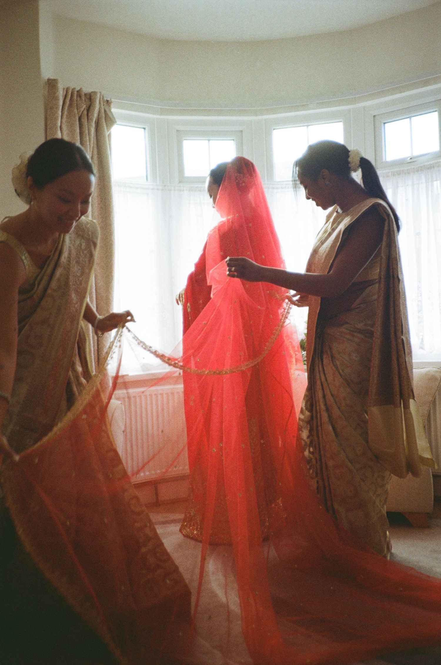South Asian bride in red veil and sari assisted by two women in gold saris during wedding preparations indoors.