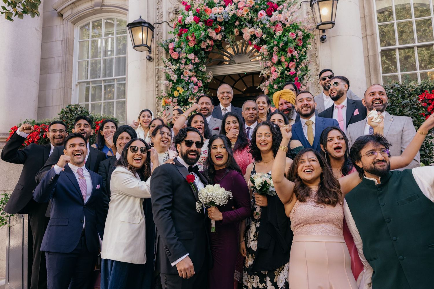 Large wedding group photo on decorated steps with floral arch, guests in colorful formal attire celebrating joyfully.