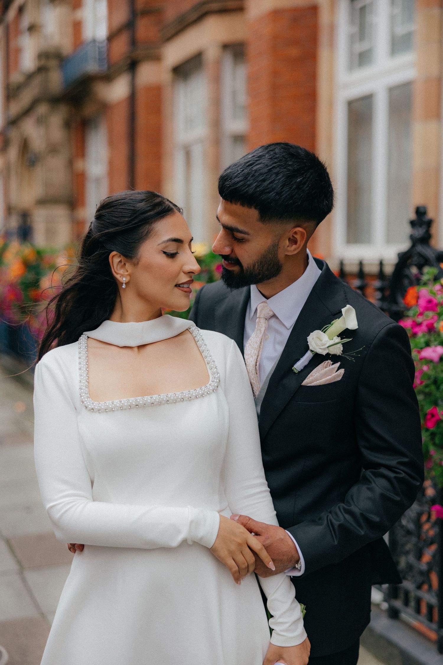 A romantic couple in wedding attire share intimate moments in front of a brick building with colorful flowers.