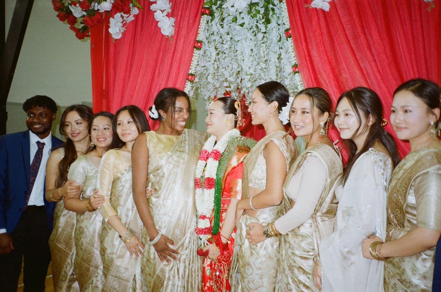 A bride in traditional attire poses with bridesmaids in gold sarees under floral decorations at a wedding ceremony.