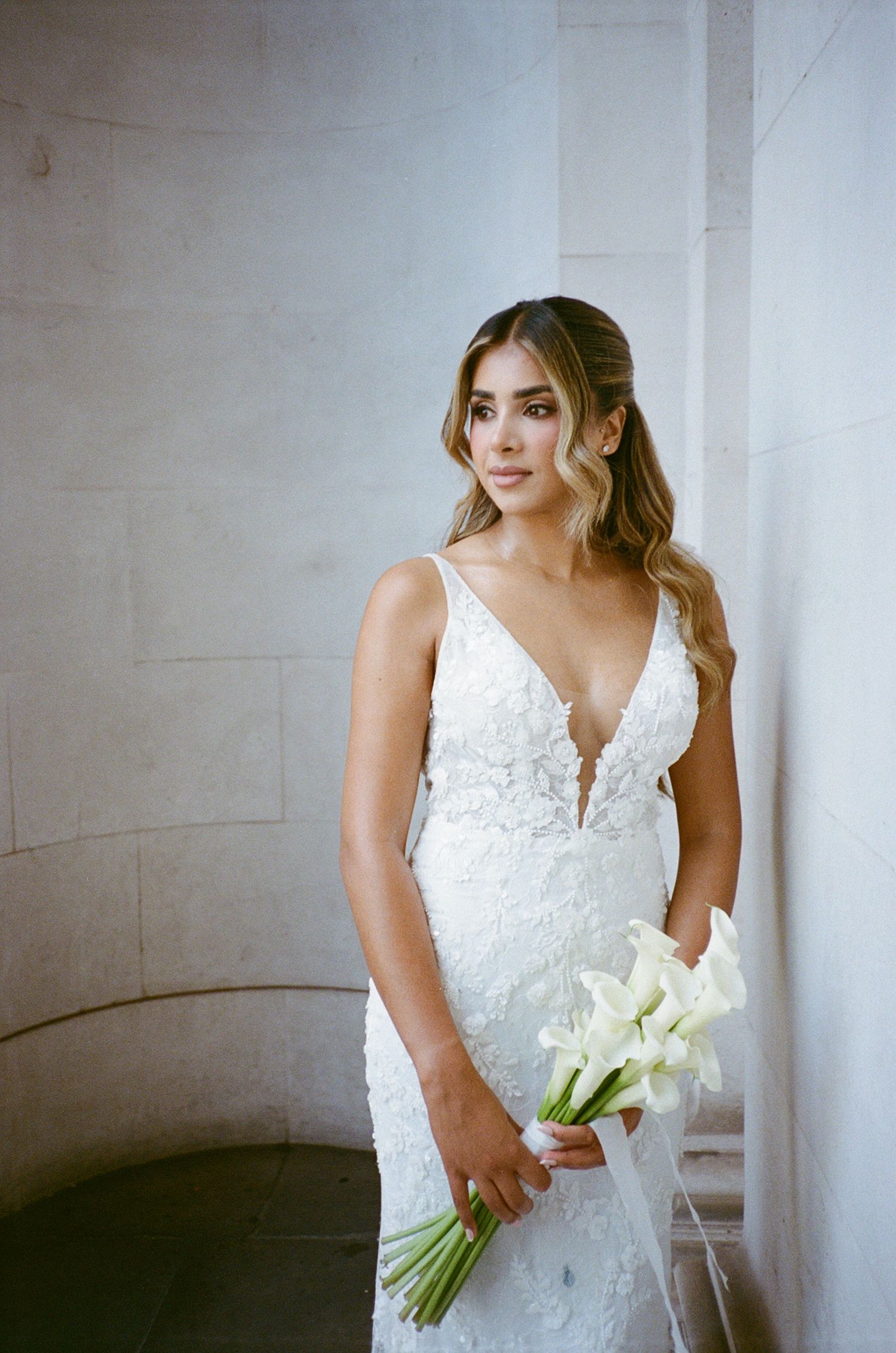 Bride in a fitted lace V-neck wedding gown holding white calla lilies, posing elegantly near stone columns.