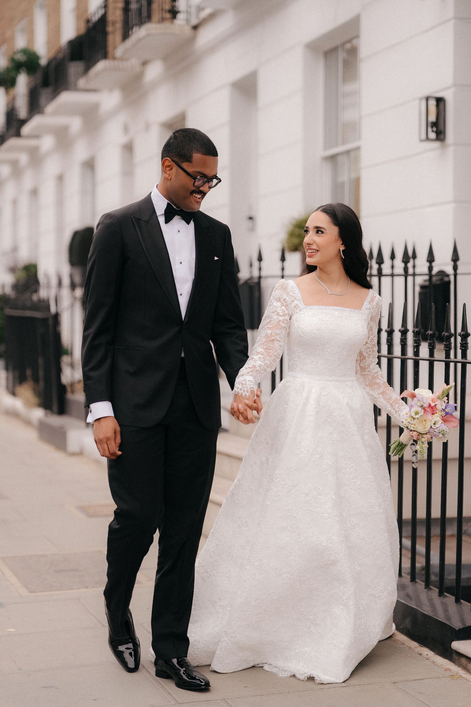 Newlyweds stroll along white urban buildings during wedding photoshoot.