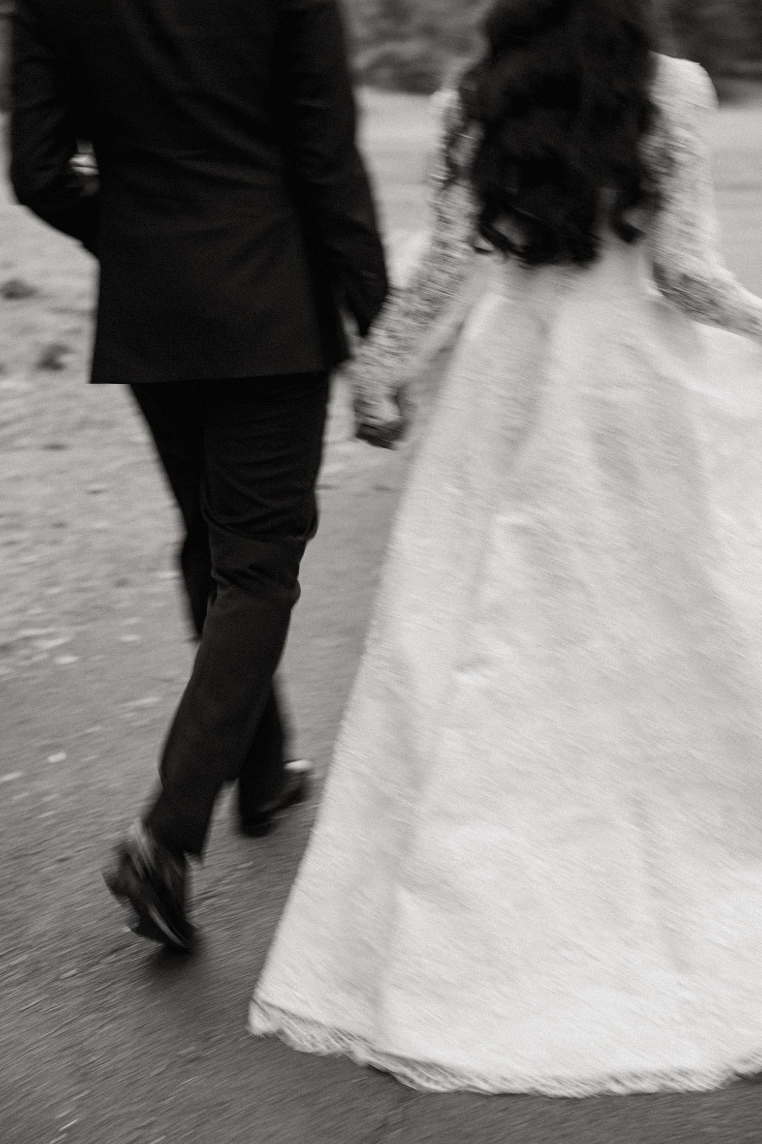 Black and white motion shot of newlyweds walking together, bride in lace gown.