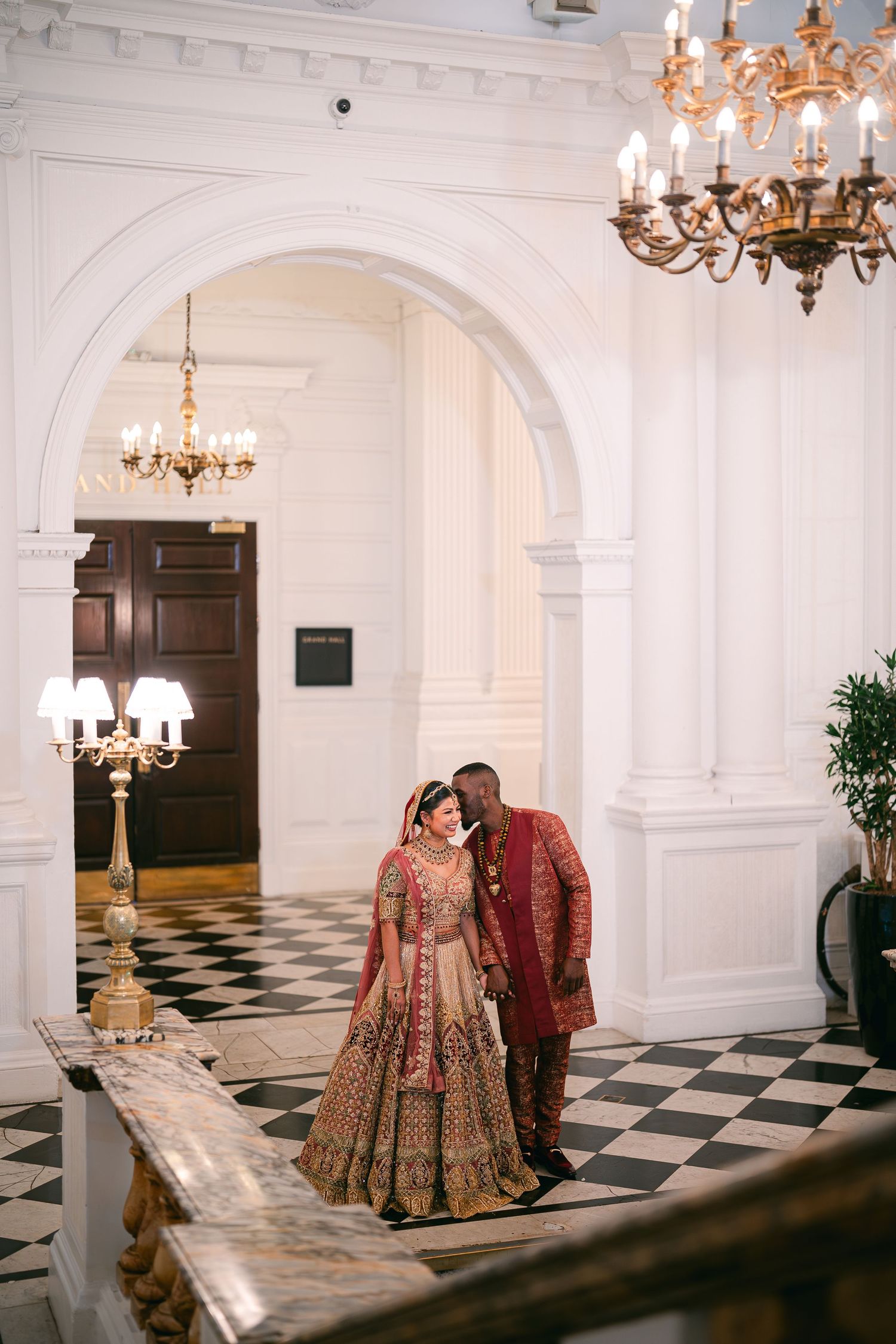 A couple in traditional Indian wedding attire poses in an elegant hall with checkerboard floors and chandeliers.