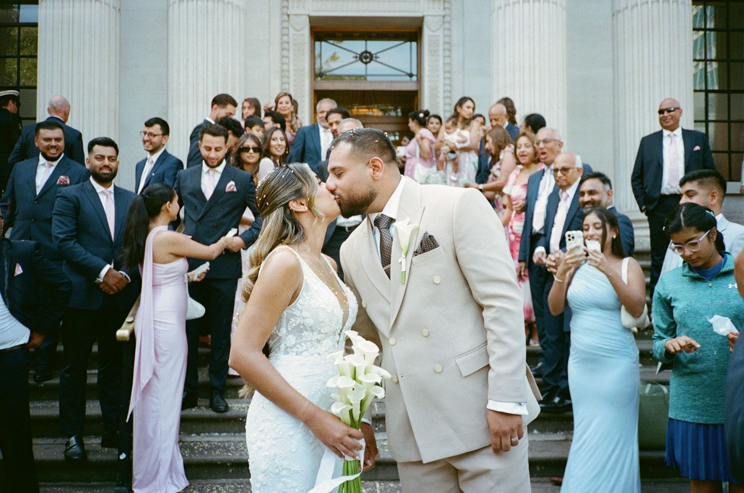 Bride and groom share a kiss on steps surrounded by wedding guests during outdoor ceremony.