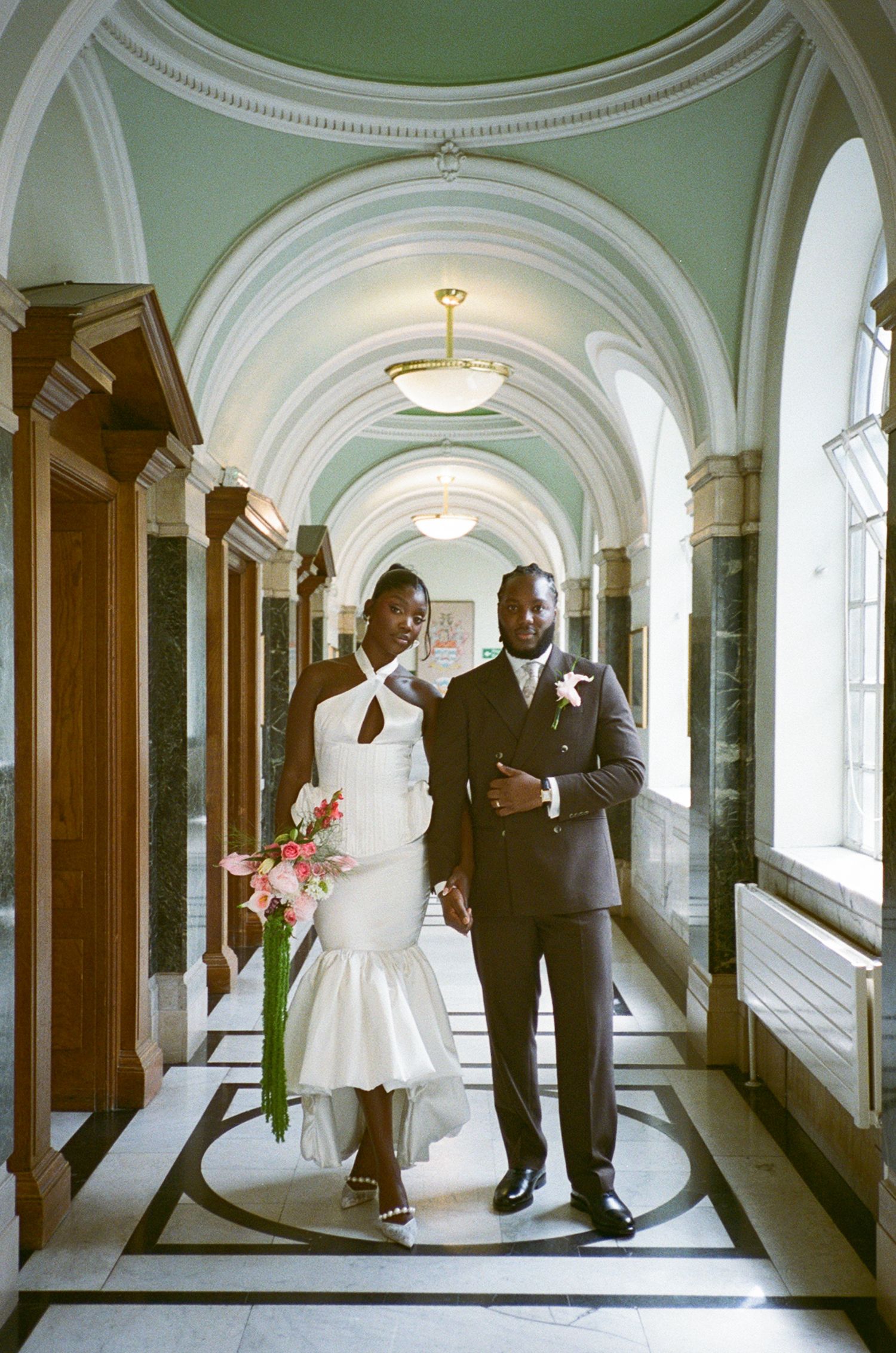 Elegant newlyweds pose in a grand arched hallway, bride in white mermaid dress holding pink florals, groom in dark suit.