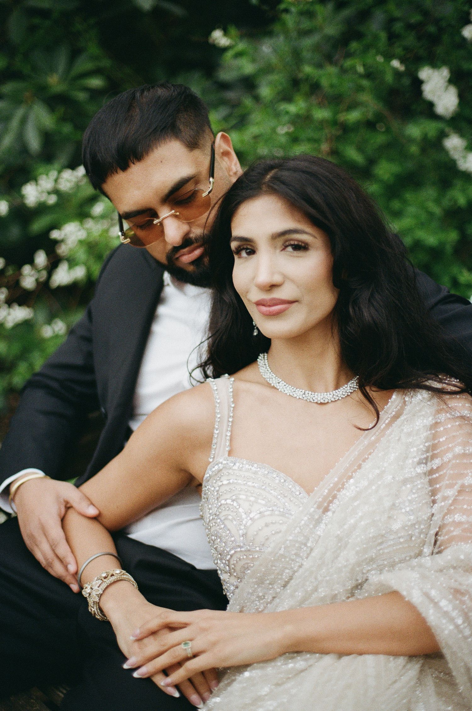 Elegant couple in wedding attire sitting outdoors, bride in beaded gown with diamond jewelry, groom in black suit.