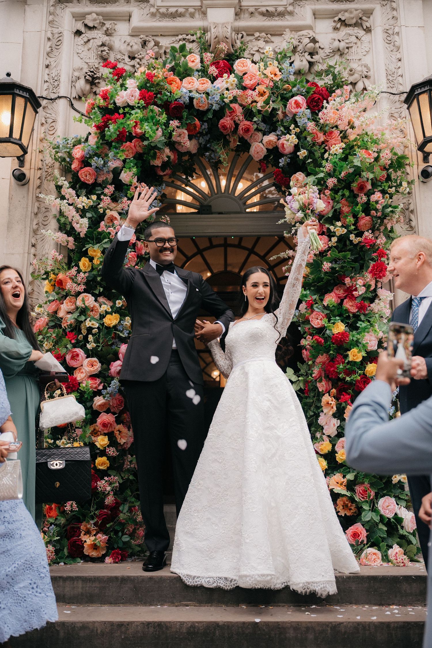 Guests celebrate as newlyweds pose under an ornate floral arch at their wedding venue.