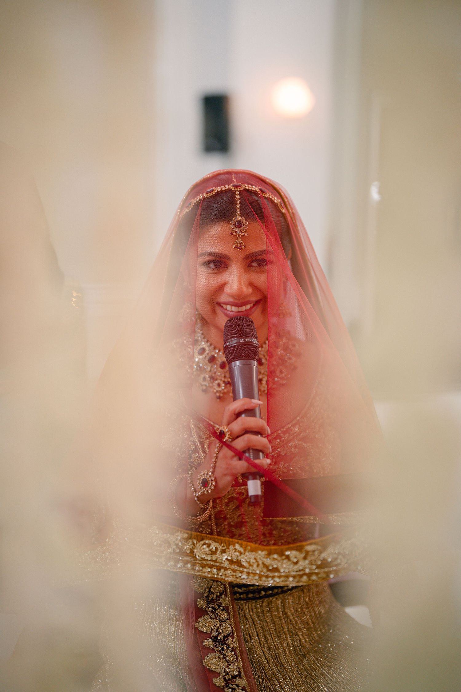 Traditional Indian bride wearing red and gold attire with intricate jewelry seen through a soft focus veil.