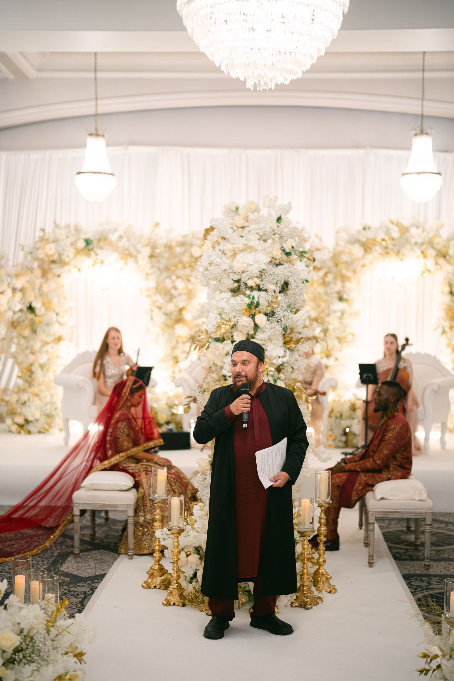 Wedding ceremony moments captured in a white decorated hall with floral arrangements and chandeliers.