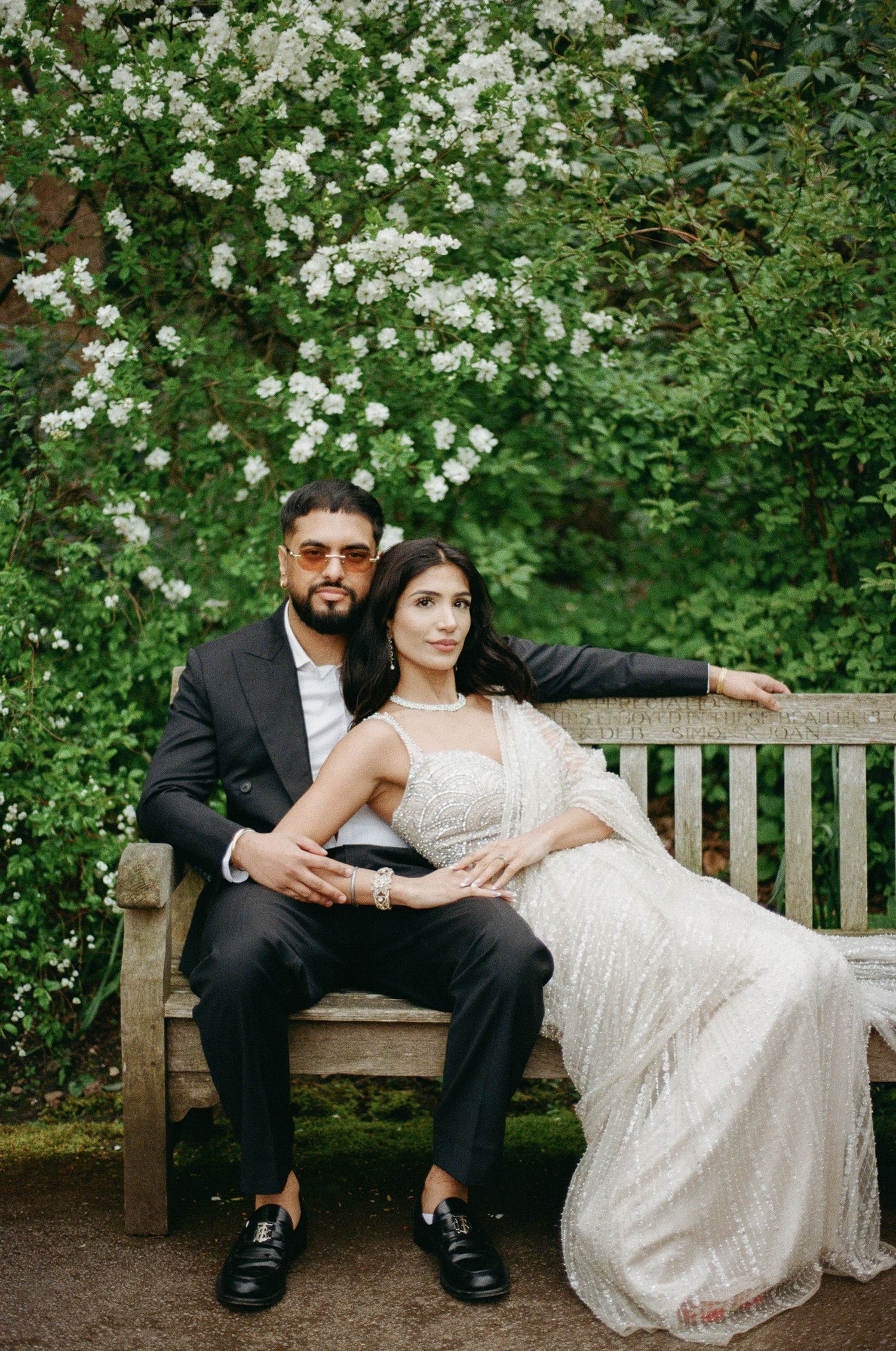 Couple in wedding attire sitting on a garden bench surrounded by blooming white flowers and lush greenery.