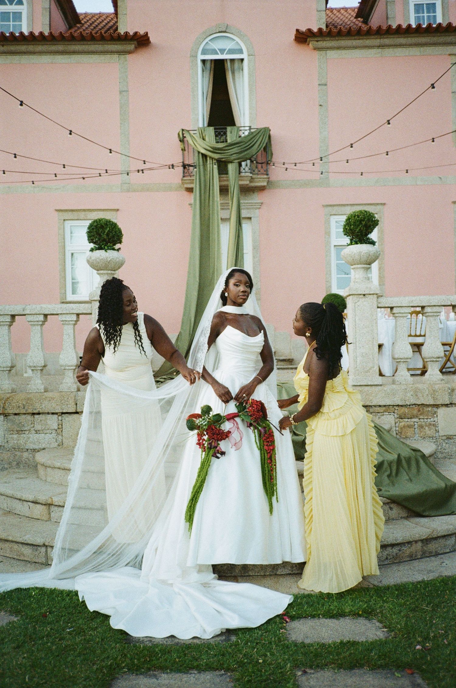 Bride in white gown with veil held by two bridesmaids outside a pink villa.