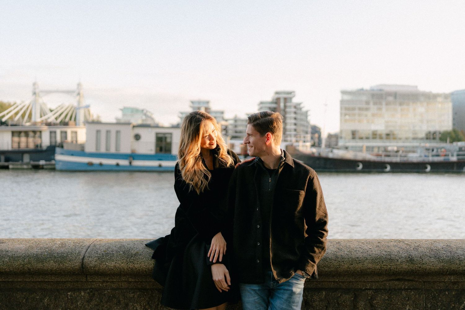 A couple stands together against a cityscape backdrop near the waterfront.