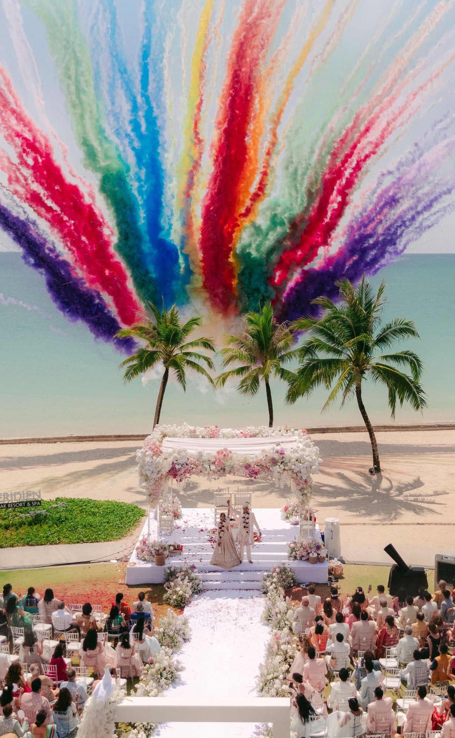 Colorful smoke effects create rainbow canopy over beachfront wedding reception with palm trees.