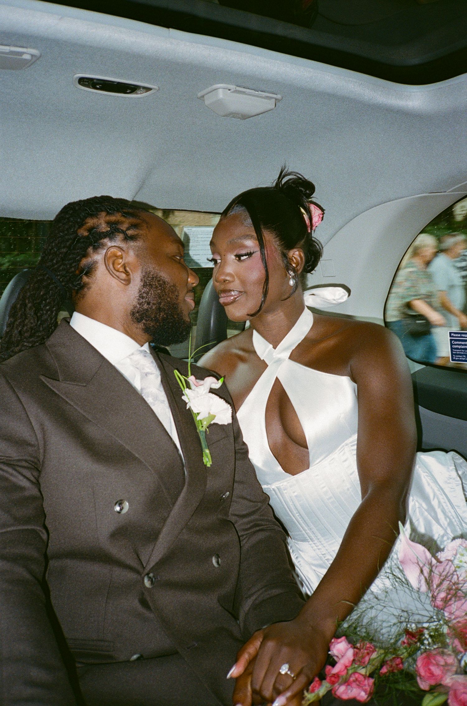 A bride in a white halter gown and groom in a dark suit share a romantic gaze inside a car surrounded by pink flowers.