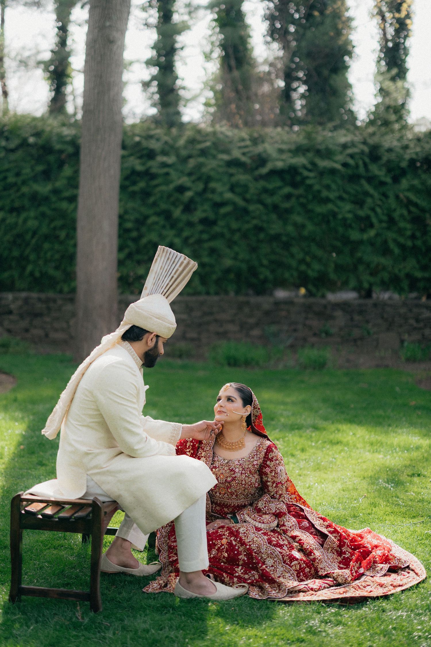 South Asian bride in red lehenga sits on grass while groom in white sherwani and turban looks at her outdoors.