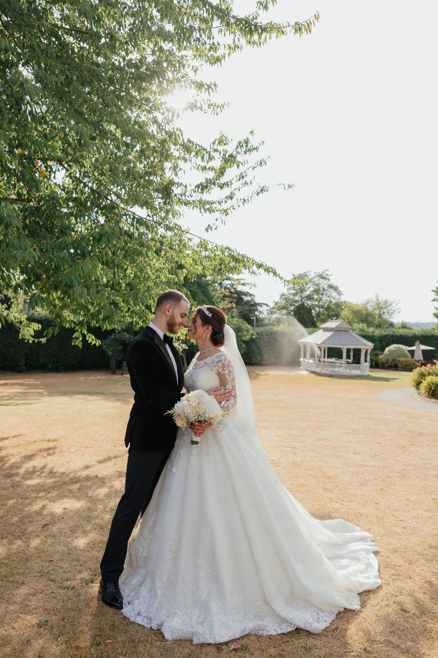 Bride and groom share a romantic moment in lush garden, bride wearing lace ballgown with veil, groom in black tuxedo.