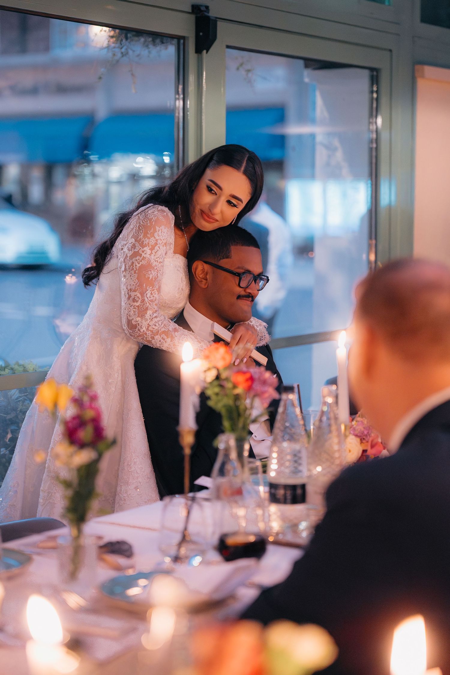 Bride in lace gown embraces groom at candlelit wedding reception dinner table with colorful floral centerpieces.