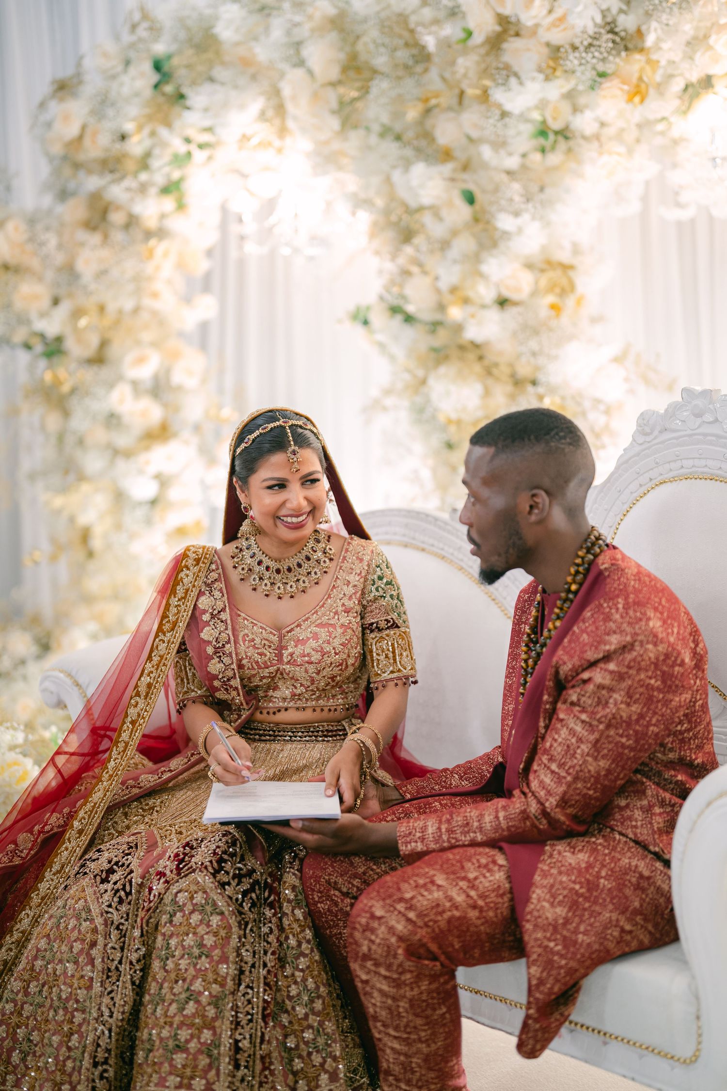 Couple in traditional South Asian wedding attire sharing a moment on ornate white sofa.