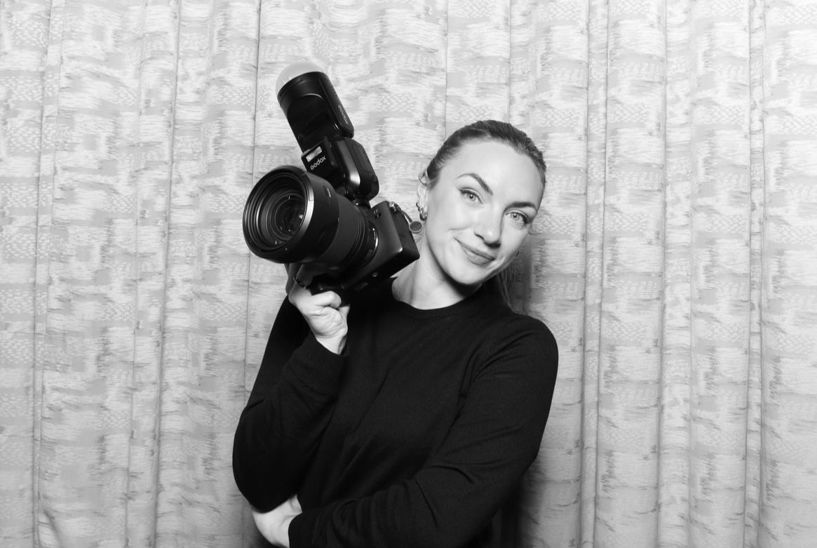 Black and white photo of someone holding a professional camera while wearing a dark sweater against a textured backdrop.