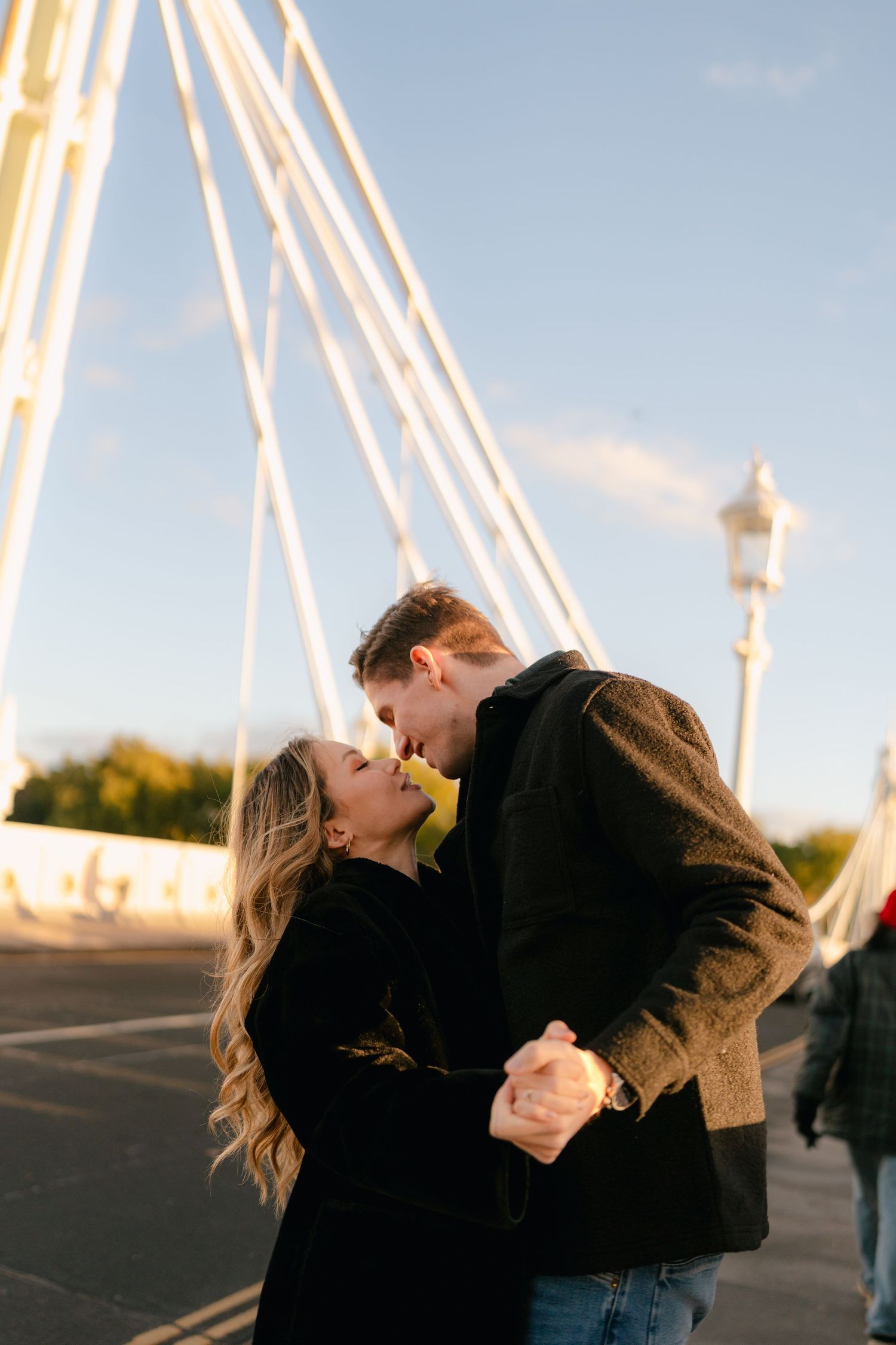 A couple embraces on a bridge with white suspension cables in the background.