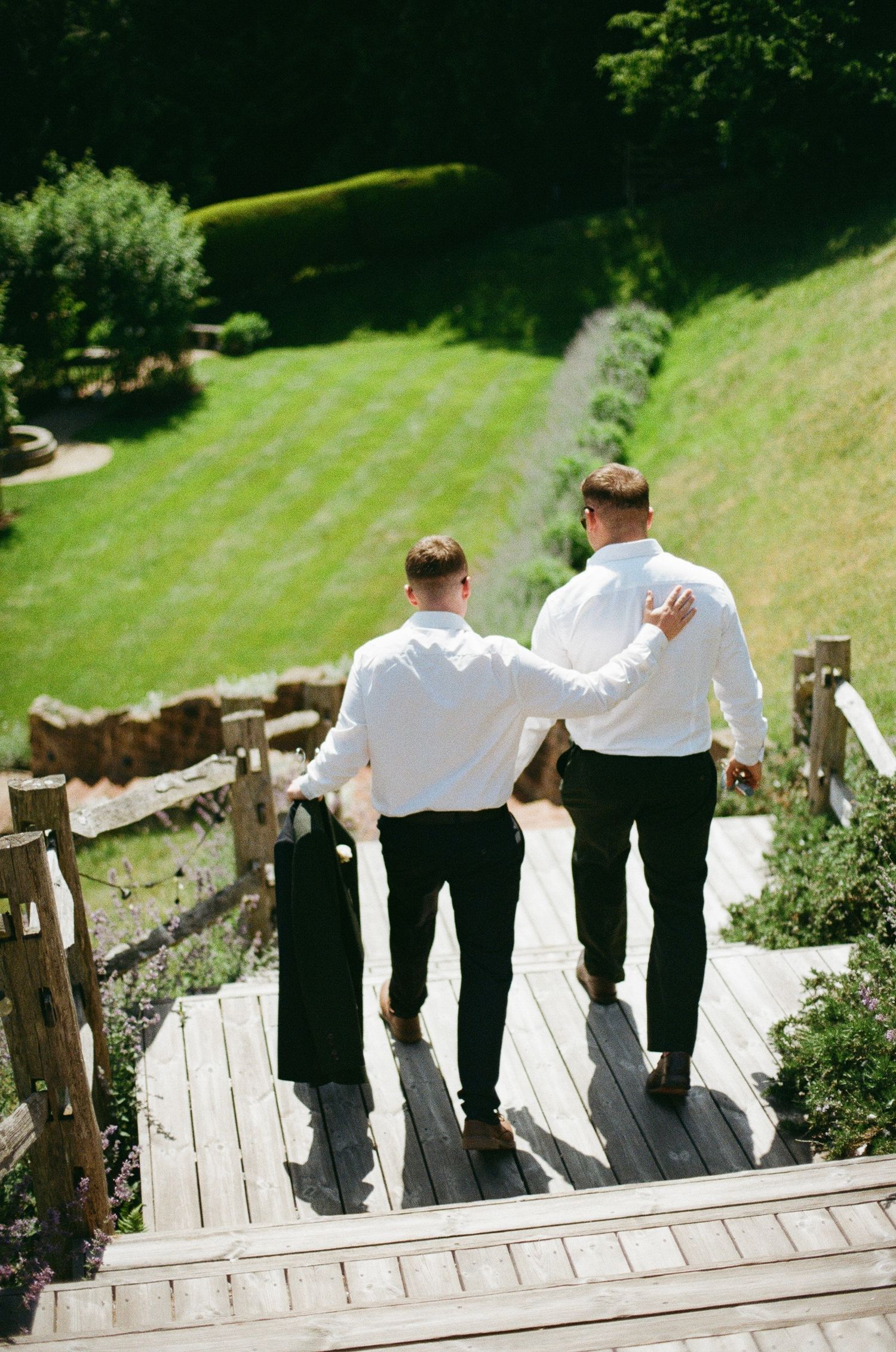 Two groomsmen in white shirts and black pants walk up wooden steps in a lush green garden setting.