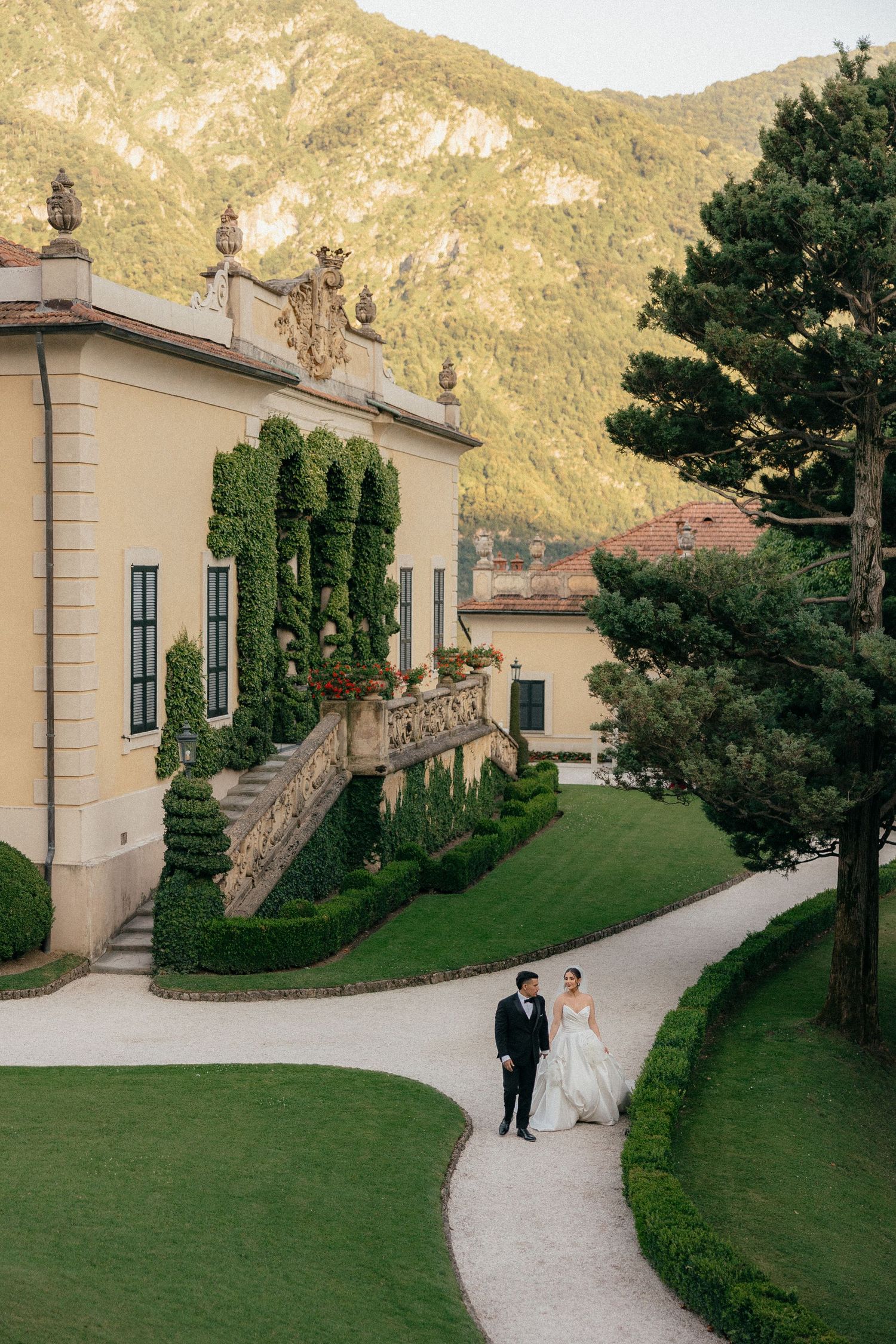 A winding garden path leads through manicured hedges and trees to an elegant villa in the Mediterranean countryside.
